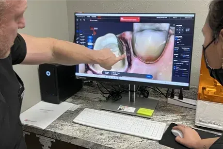 Dentist points at digital tooth scan on monitor with patient observing in office.