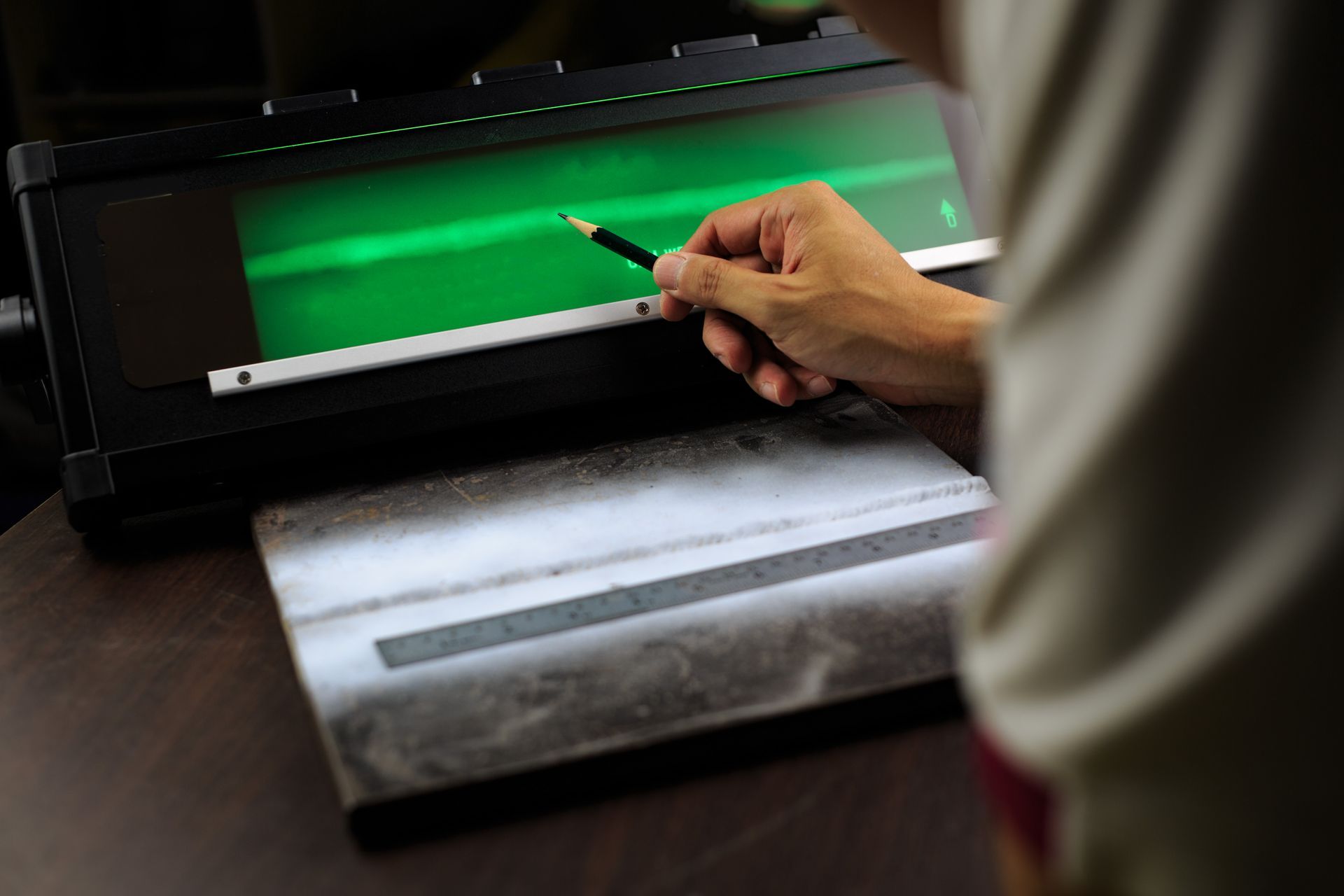 Person examining a green fluorescent line on a screen with a pencil, possibly for quality control of a metal part.