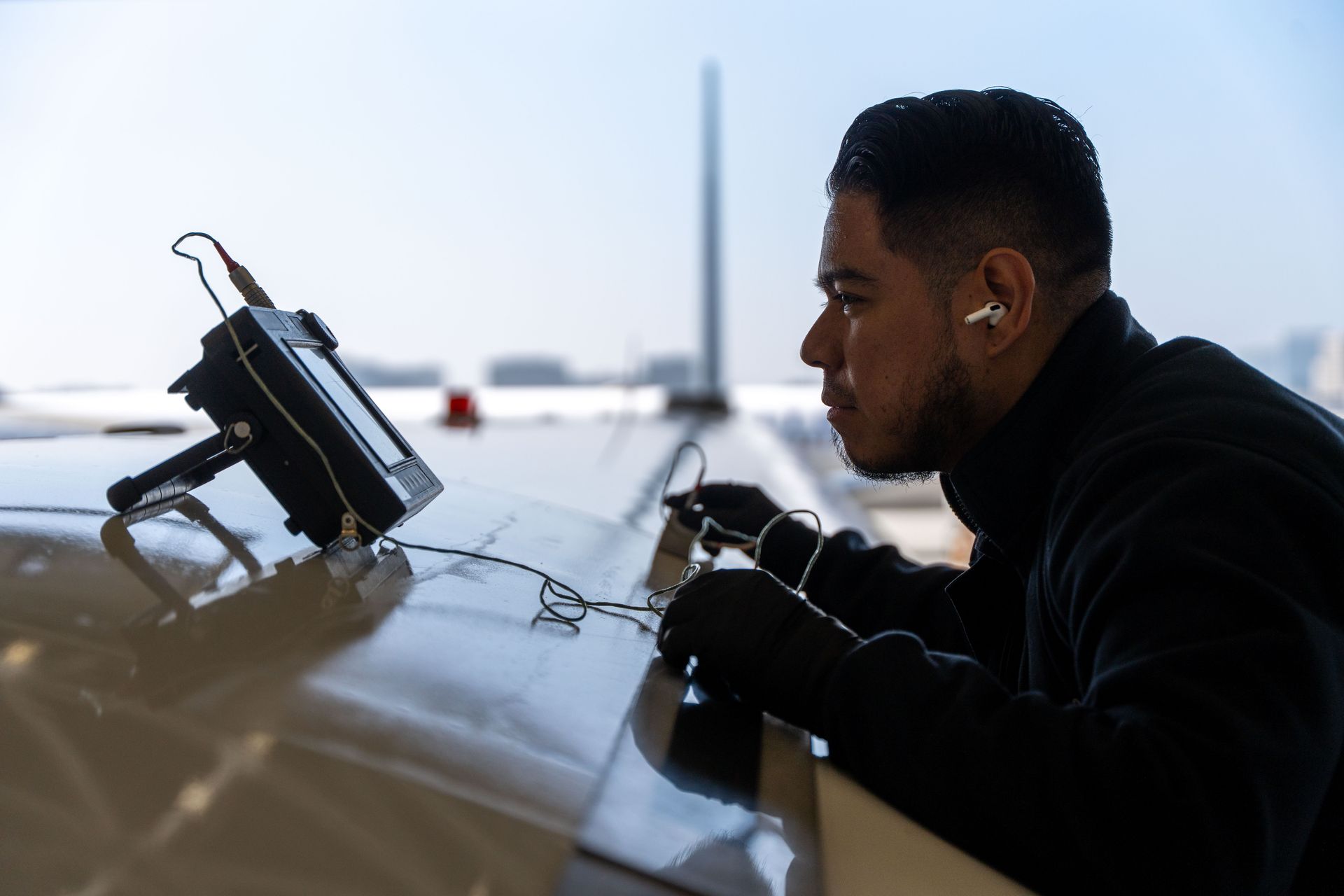 A person in dark clothing works on an aircraft wing, using a small handheld electronic device connected by wires.