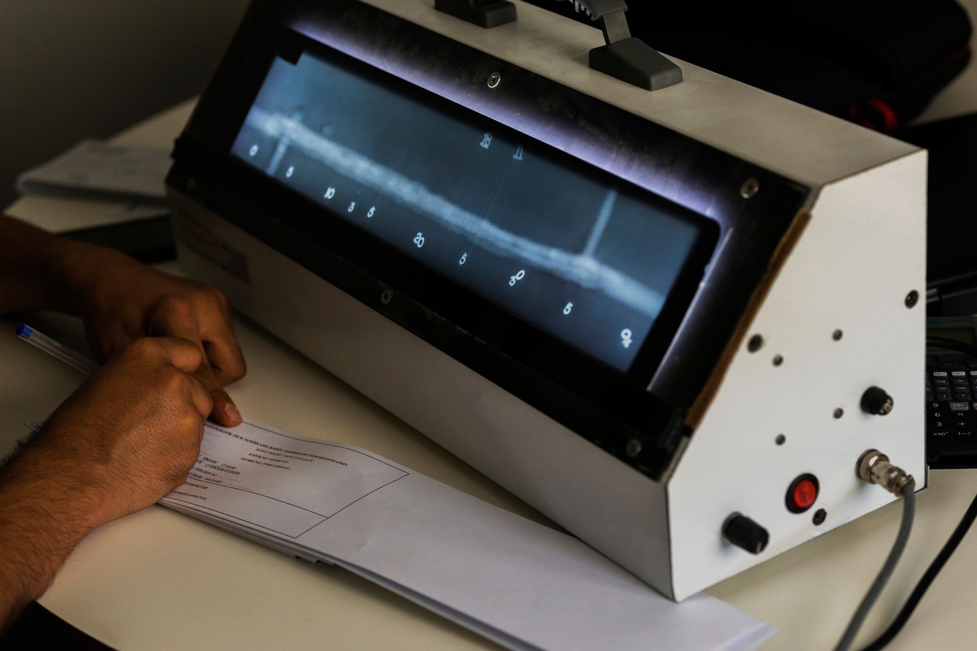 Person examining a radiographic image on a machine, with paperwork and a pen on the table.