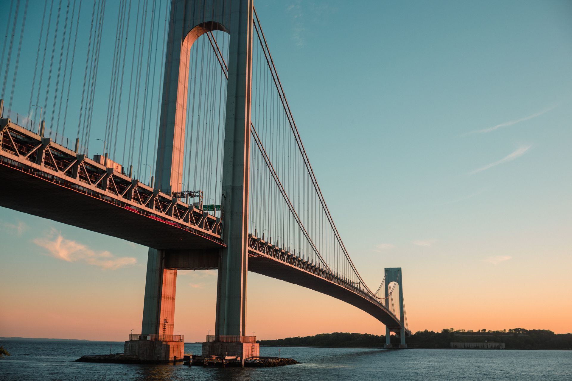 Suspension bridge at sunset over calm water, with orange-lit towers and cables against a blue sky