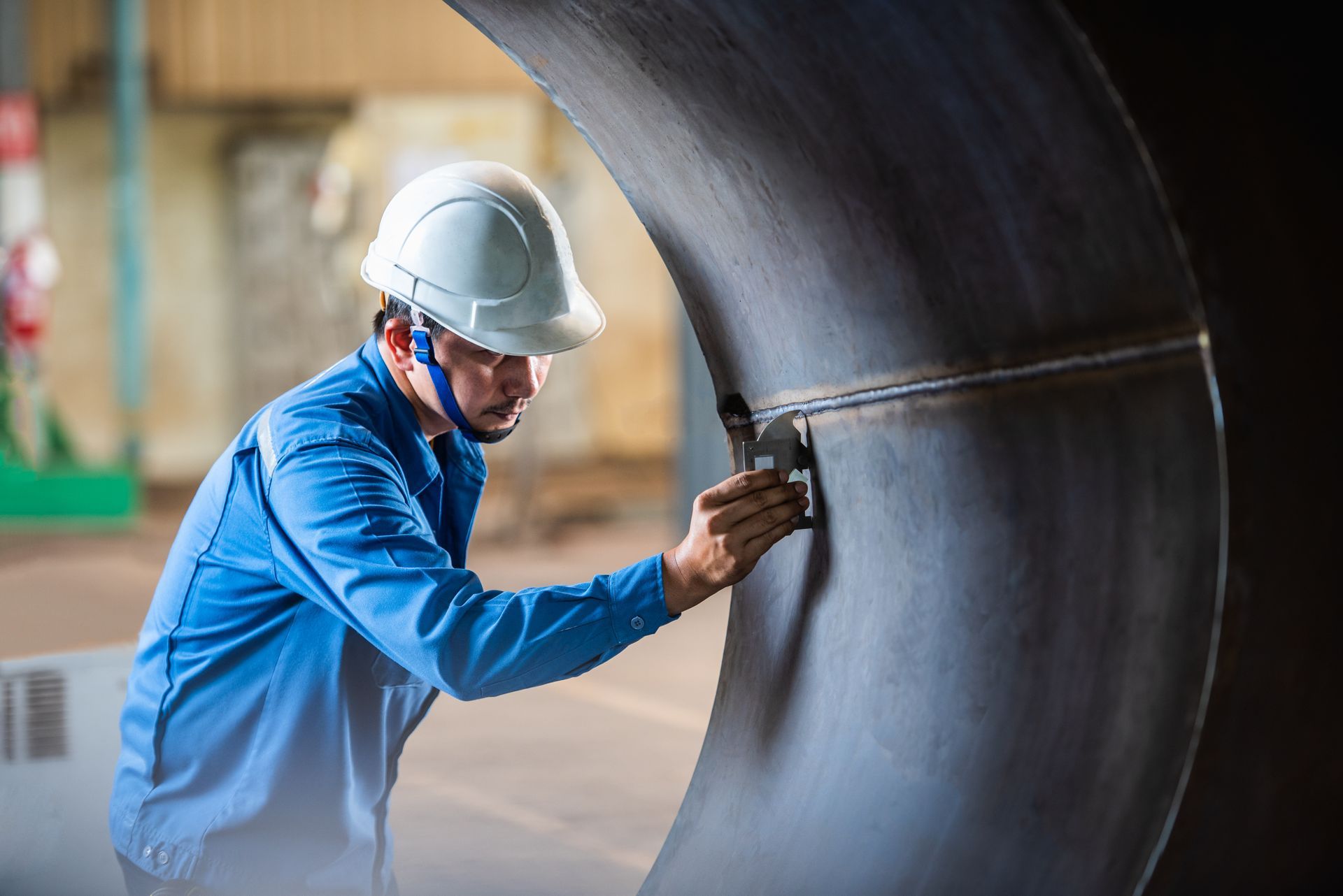 Worker in blue overalls and white helmet inspecting curved metal pipe with a tool.