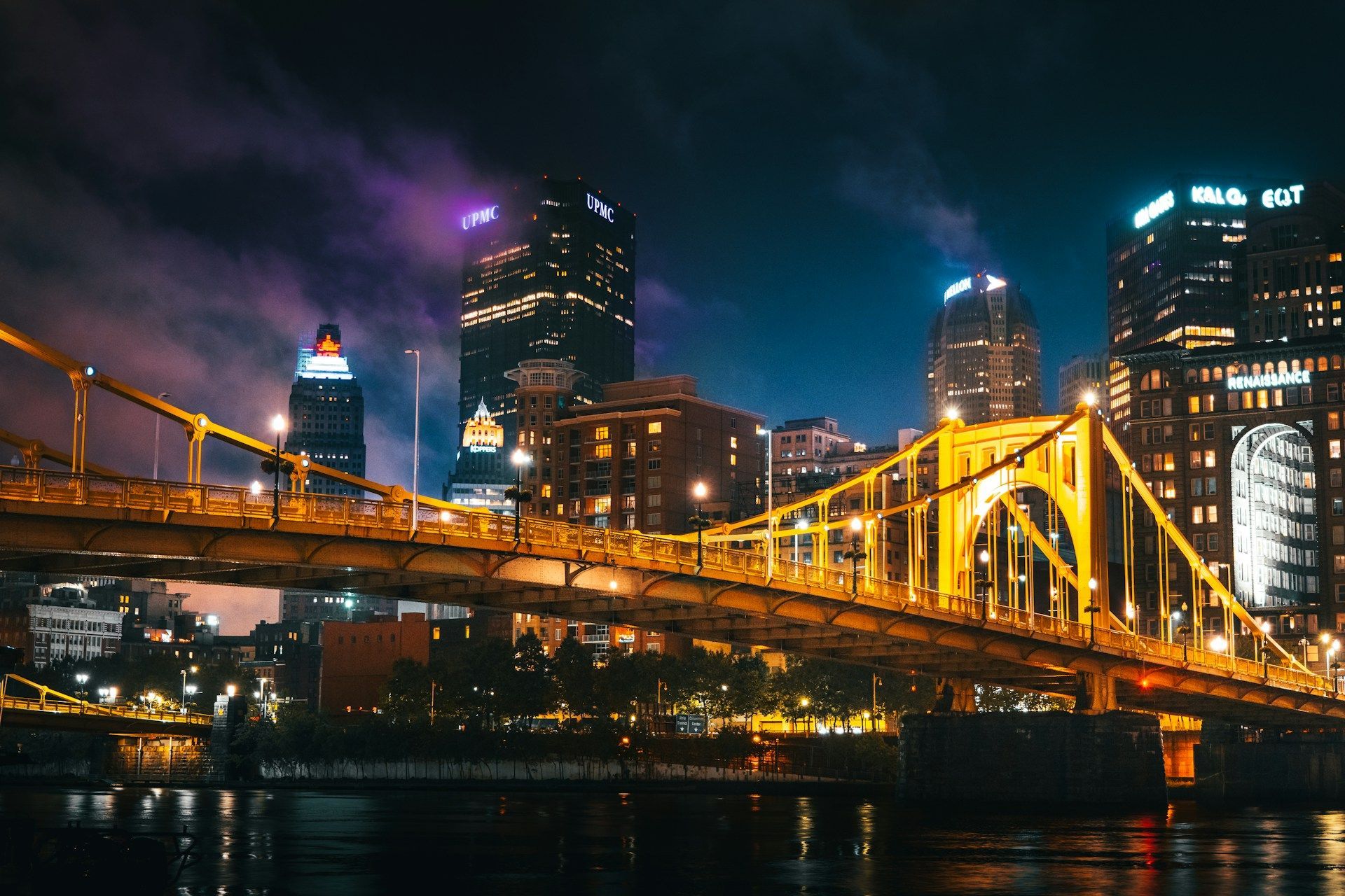 Night city skyline with a lit yellow bridge reflected on dark water at dusk in Pittsburgh, PA.