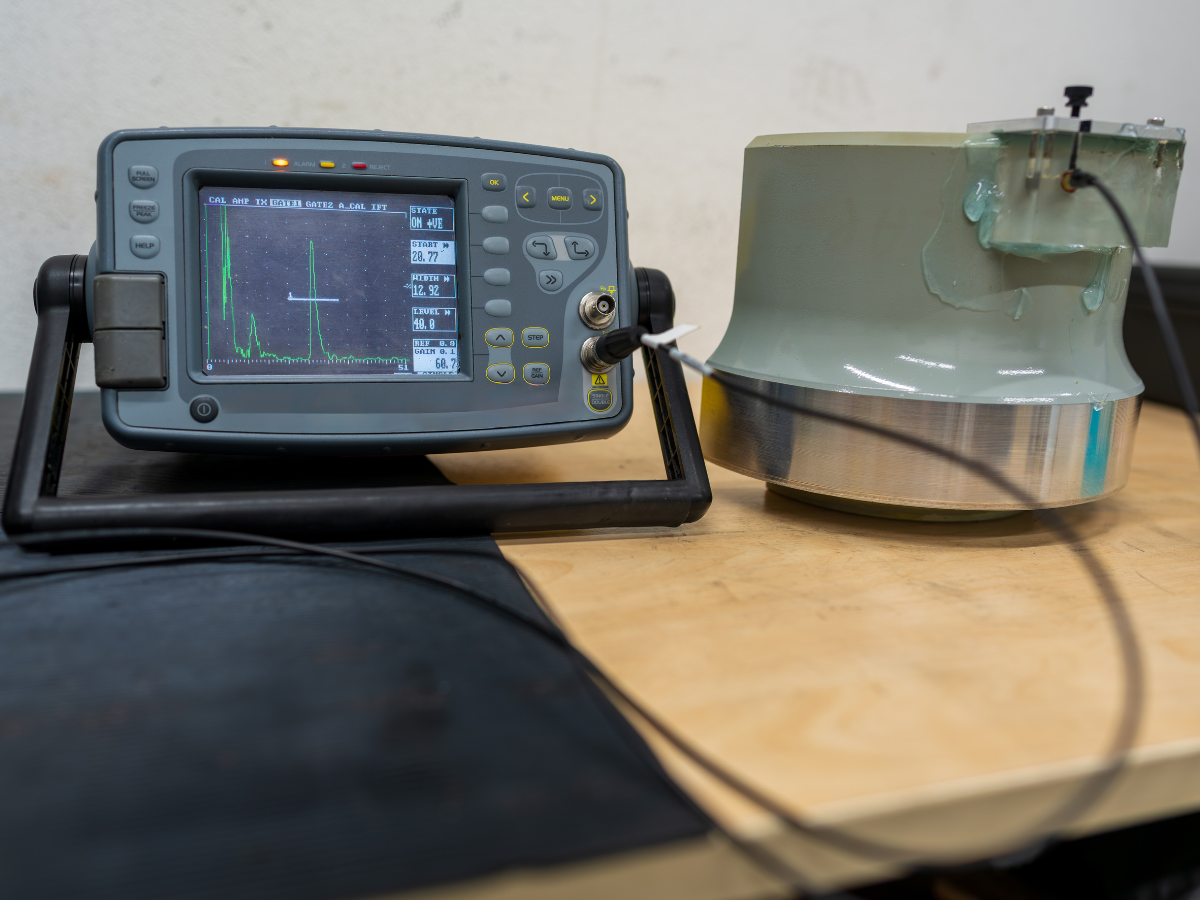 Ultrasonic testing device next to a cylindrical metal component; a green display shows a waveform.