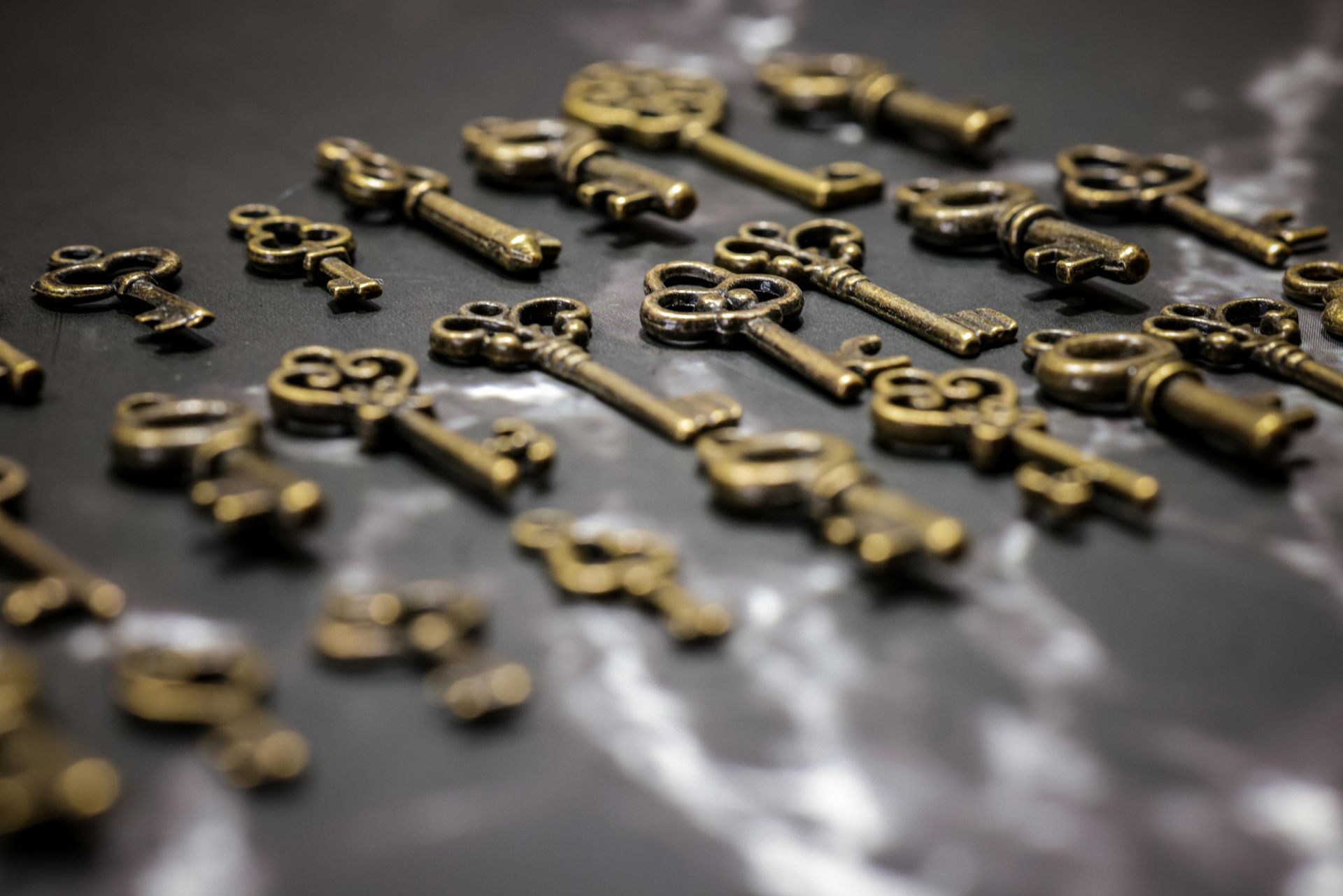 Small, antique-style keys in various sizes and designs, scattered on a dark, textured surface.