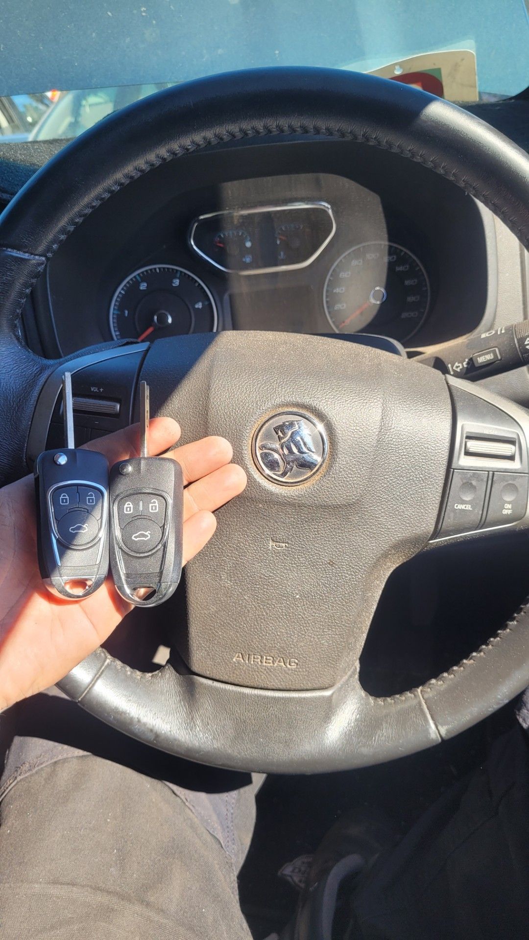 Hand Holding Two Car Key Fobs in Front of a Steering Wheel — Shaine Hunter Locksmiths in Doomadgee, QLD