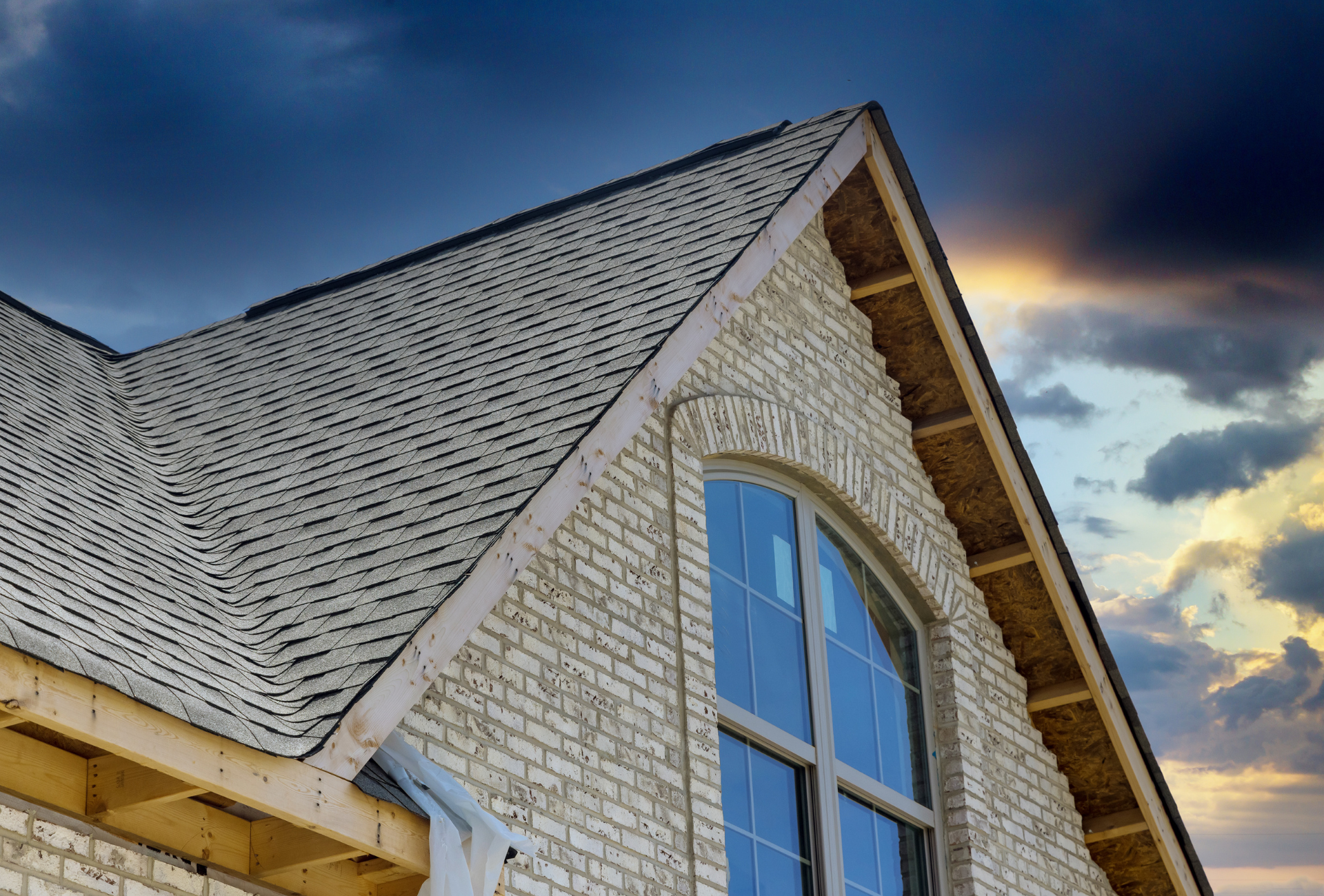 A brick building with a roof and a window with a cloudy sky in the background.