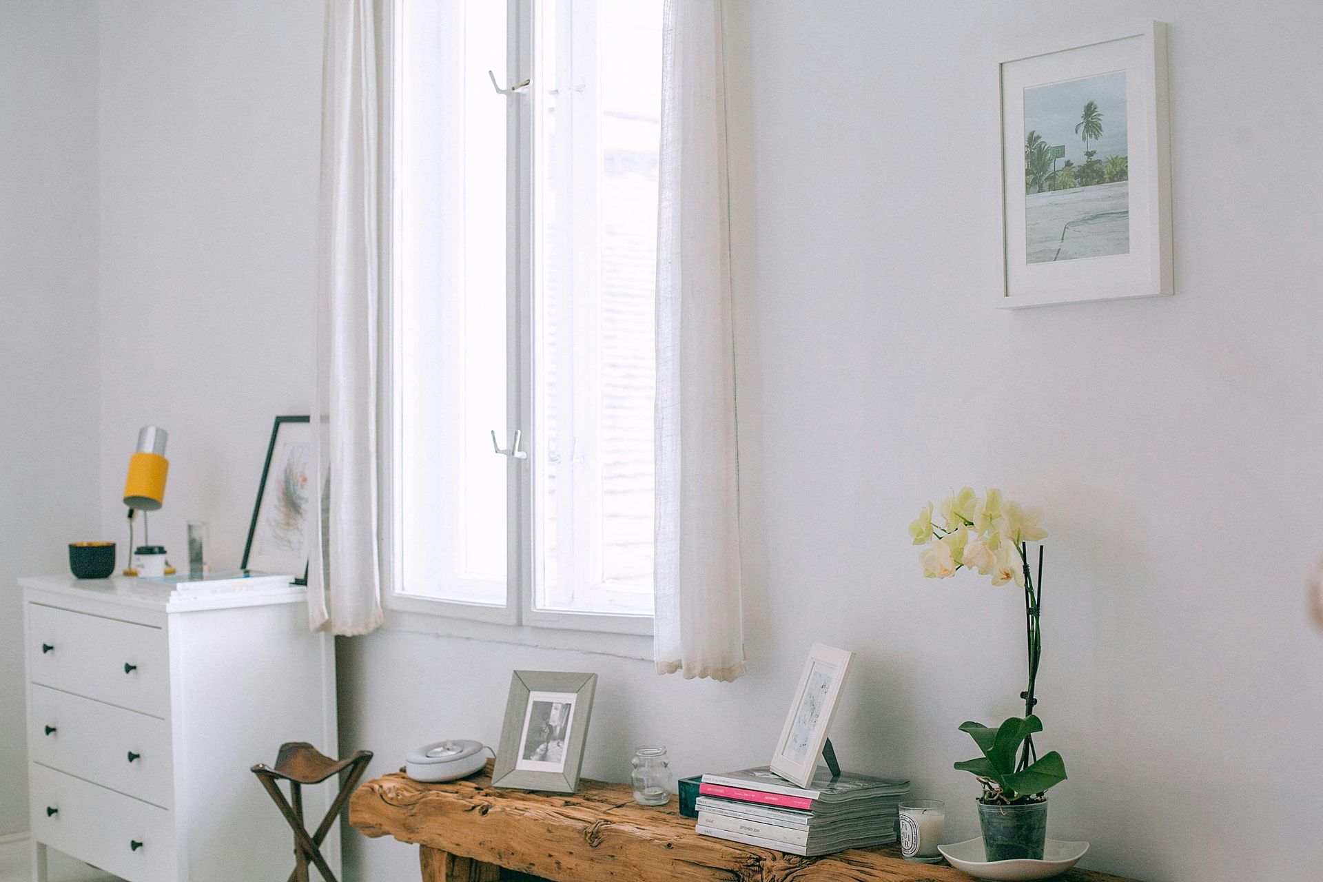 White room with window, wooden desk holding orchid, framed photos, and a white dresser.
