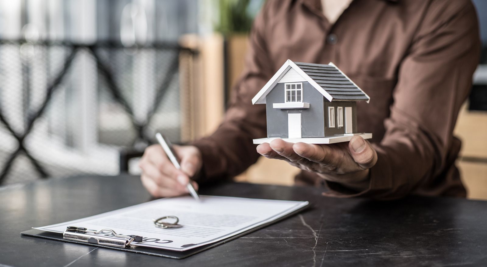 Hands holding a small house model beside a clipboard on a desk, suggesting a home sale or insurance review