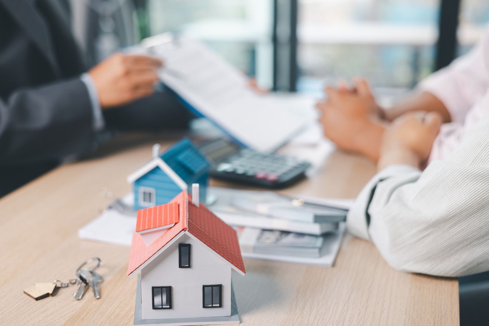 Miniature house model on a desk with blurred people discussing real estate paperwork