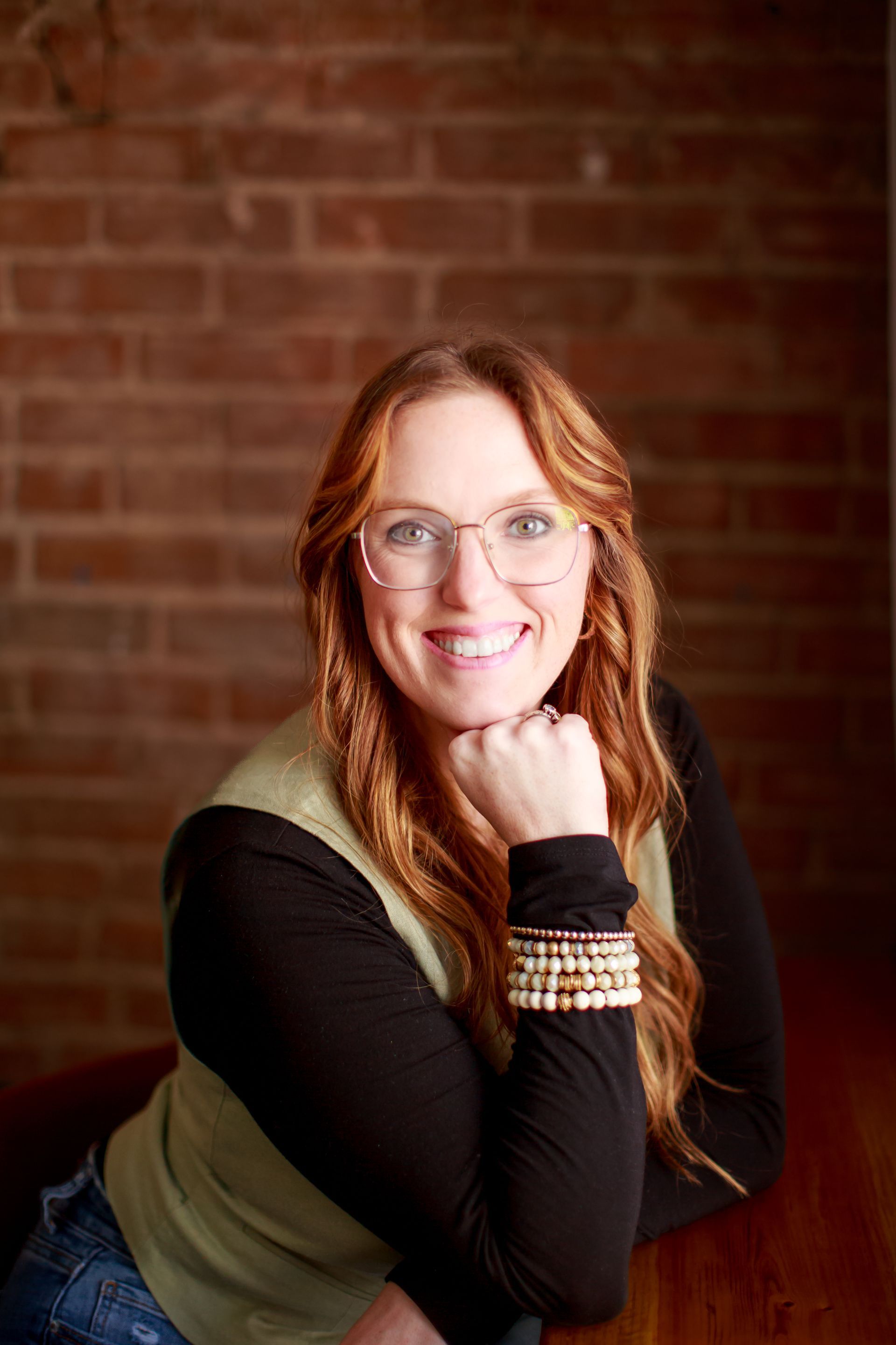 Woman with auburn hair smiles, resting chin on hand. She wears glasses, a green vest, and sits near a brick wall.