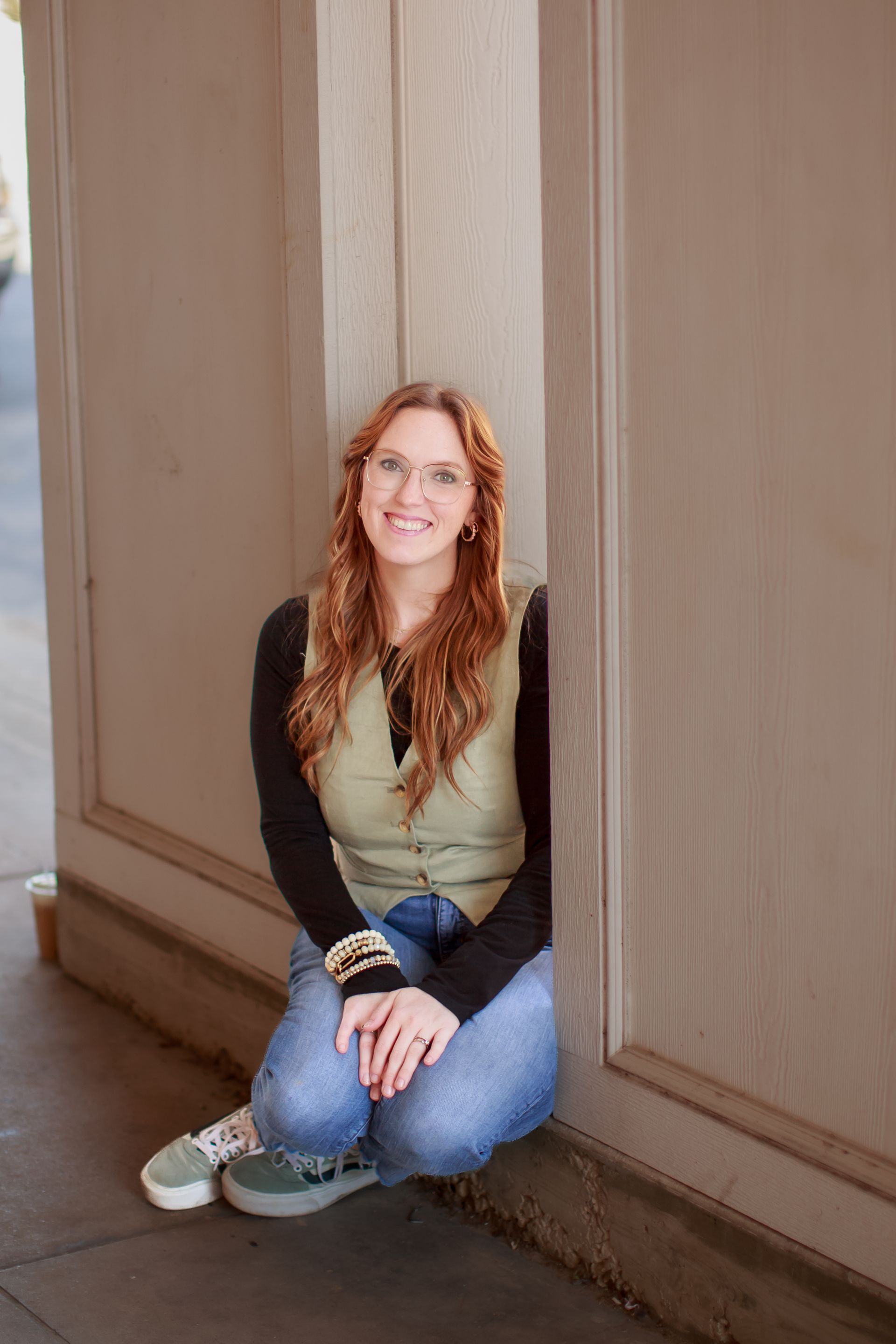 Woman kneeling by a light-colored pillar, smiling, wearing glasses, vest, jeans, and sneakers.