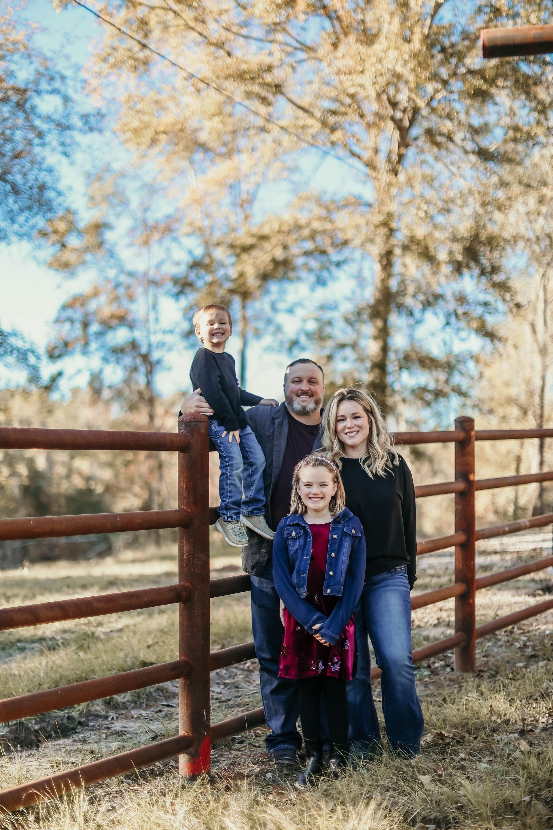 Family poses together outdoors by a wooden fence; son perched on the railing, sunny day.