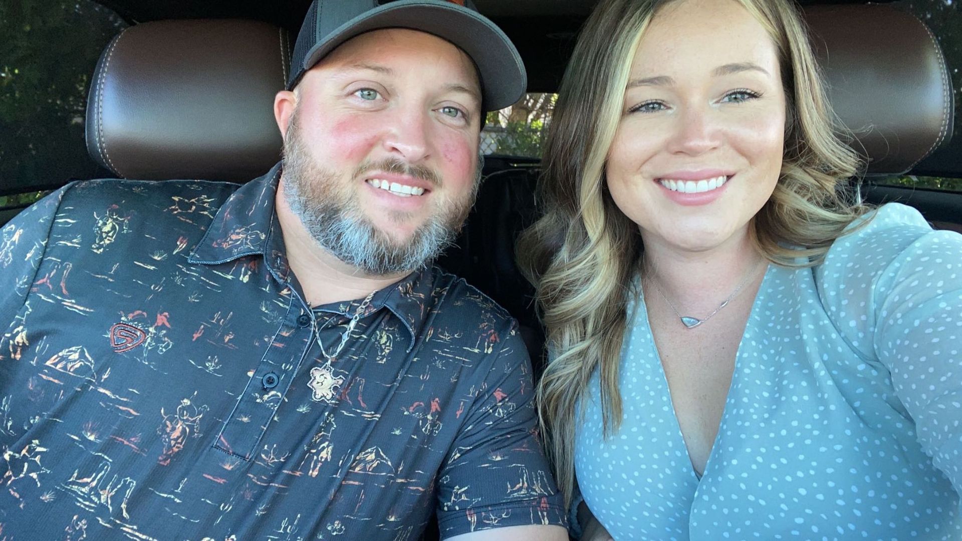 Smiling couple in car, man wearing hat and patterned shirt, woman in light blue dress.