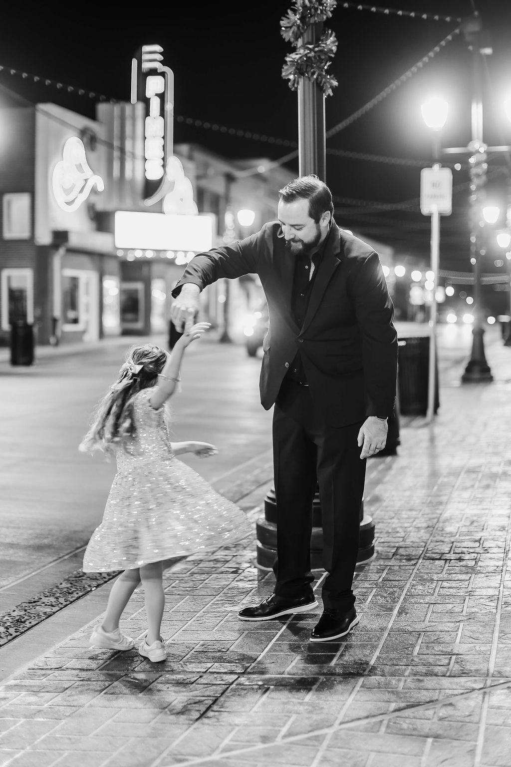 Man and young girl dancing on a brick sidewalk at night, near streetlights and a theater.