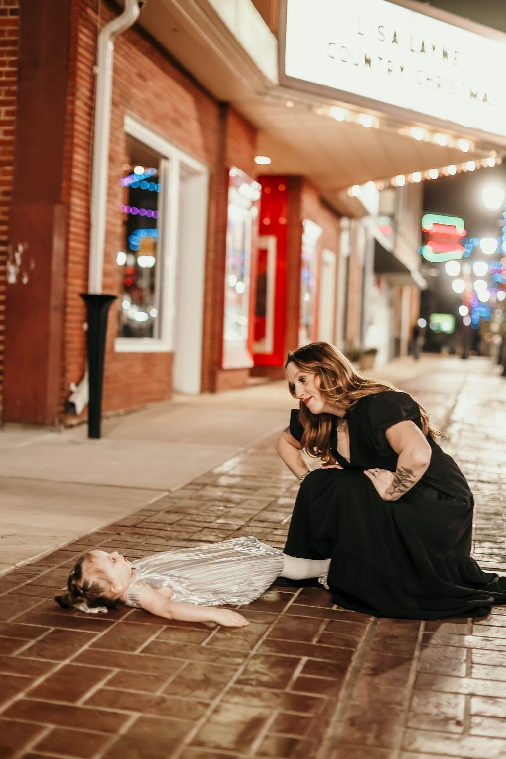 Woman kneels by a person lying on brick sidewalk, looking down. Storefront lights illuminate the scene at night.