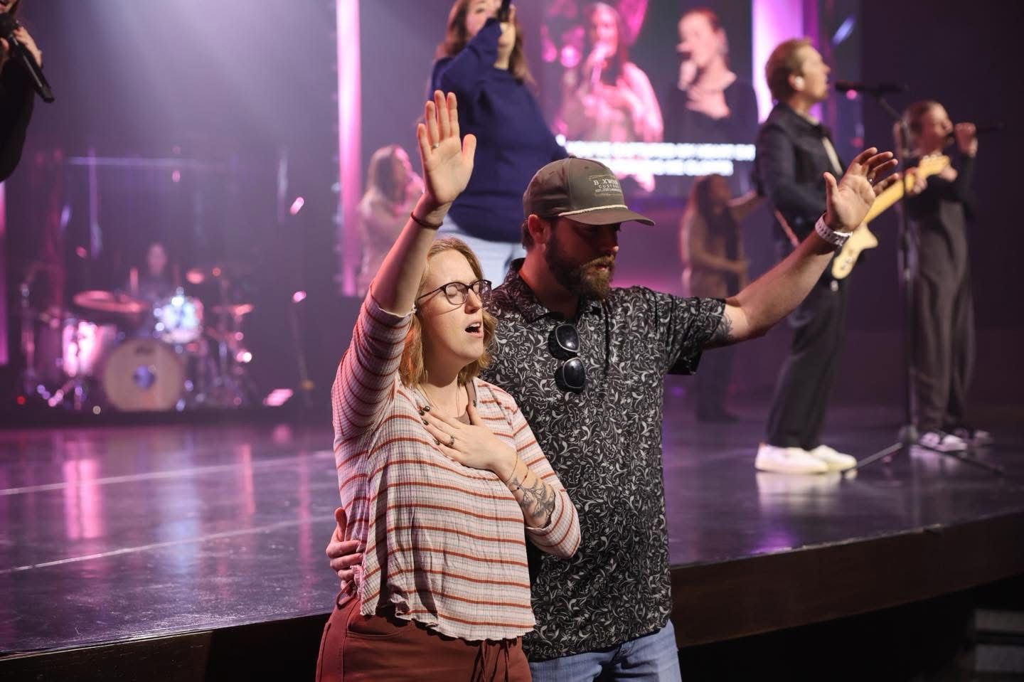 People on stage with arms raised during a performance. Stage lit with purple lights.