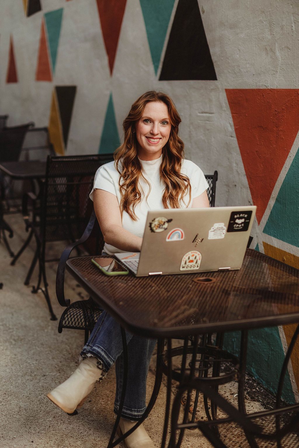 Woman with red hair sits at outdoor table, laptop open, smiling. Colorful geometric wall in background.
