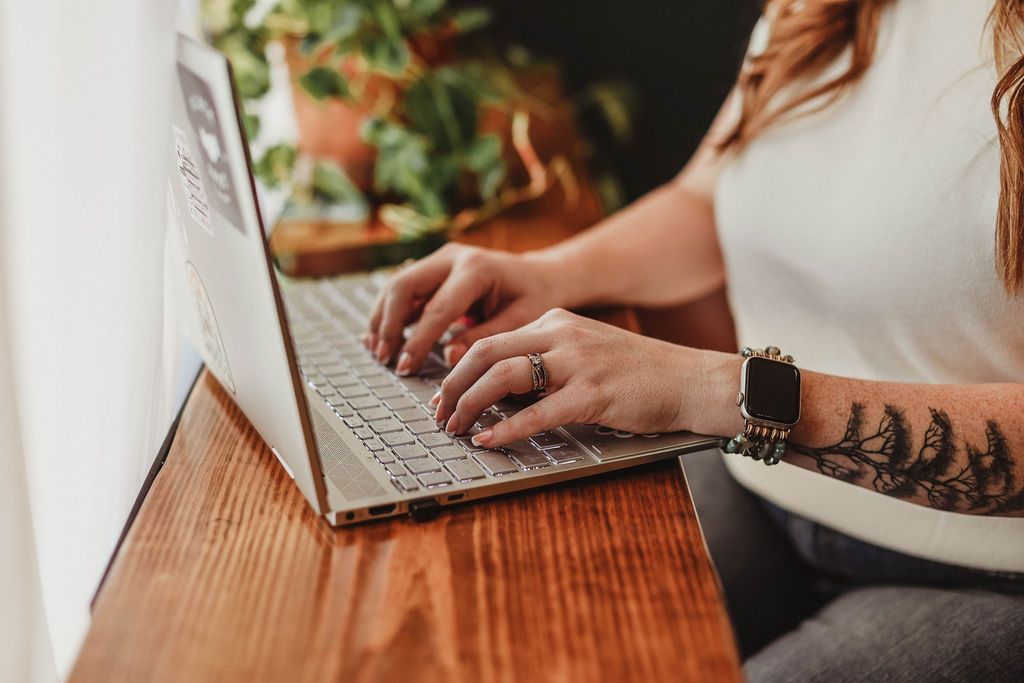 Person typing on a laptop at a wooden desk, arm tattoo visible, houseplant in the background.