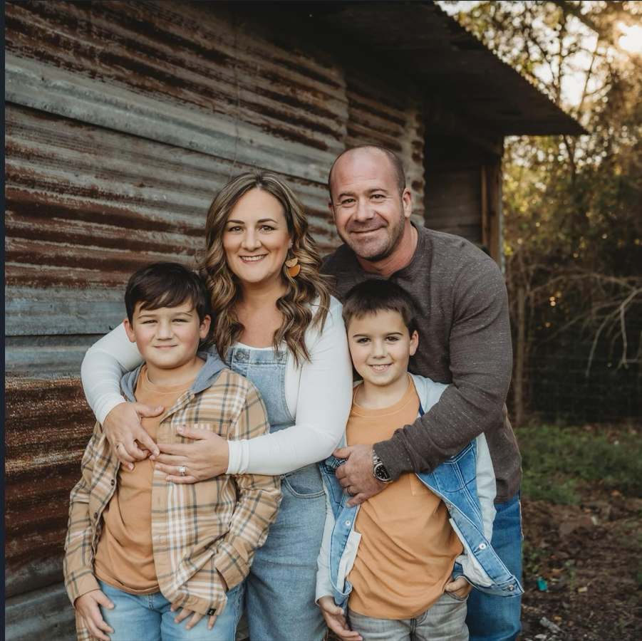 Family of four poses near a wooden fence outdoors on a sunny day.