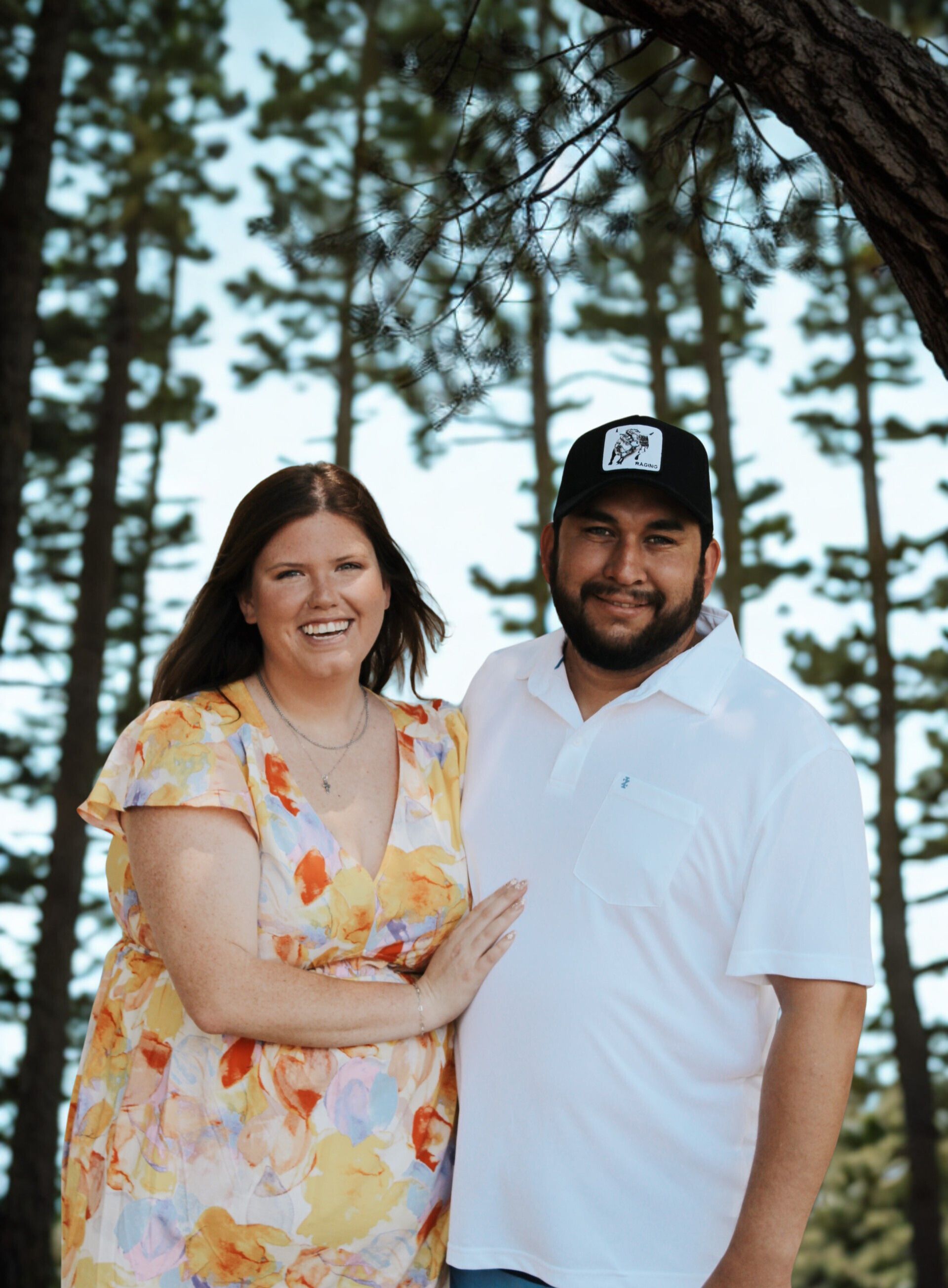 Woman in floral dress and man in a white polo, smiling in front of a forest of tall trees.