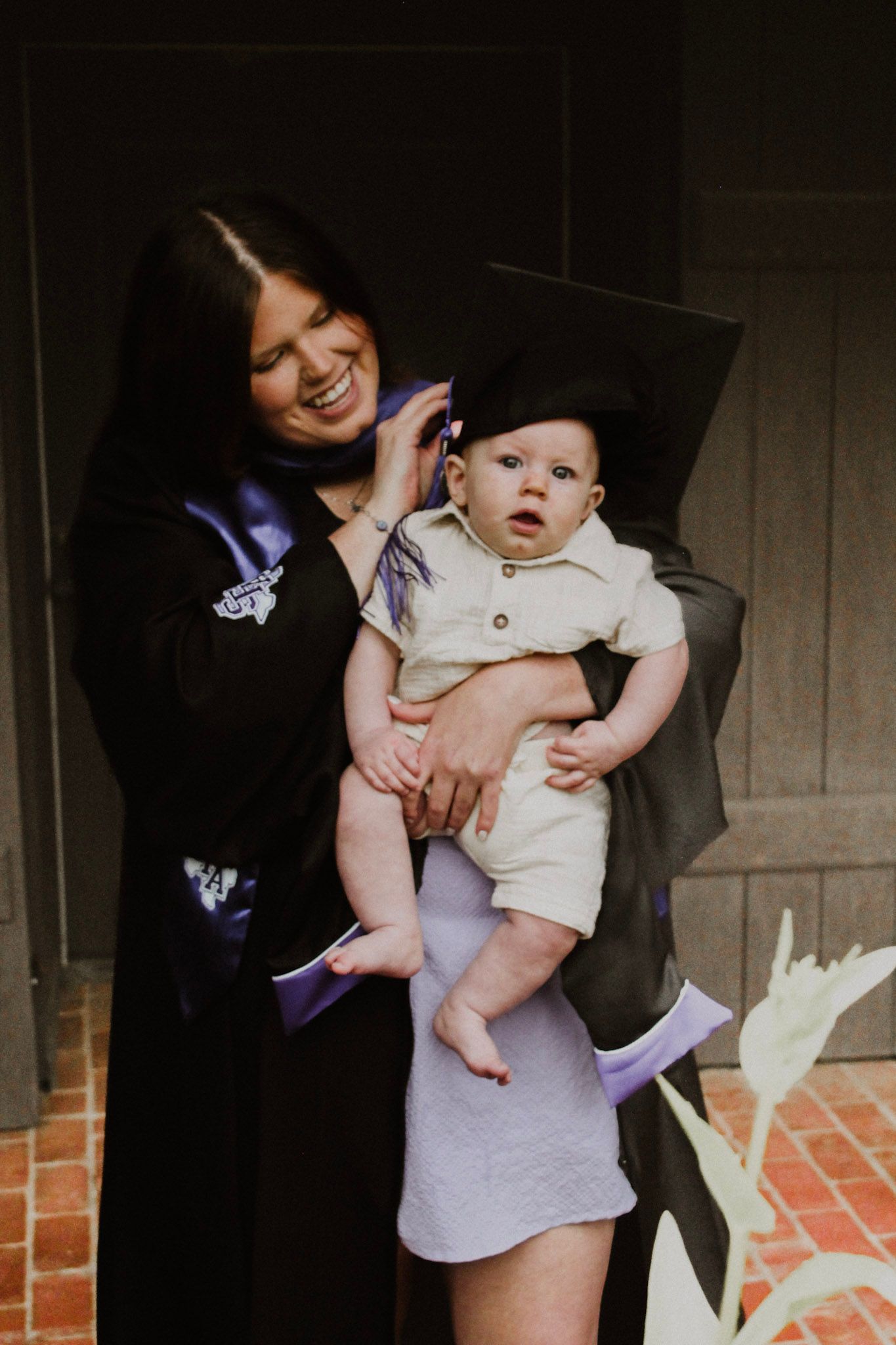 Woman in graduation gown holding a baby with a tiny graduation cap, both smiling in front of a building.