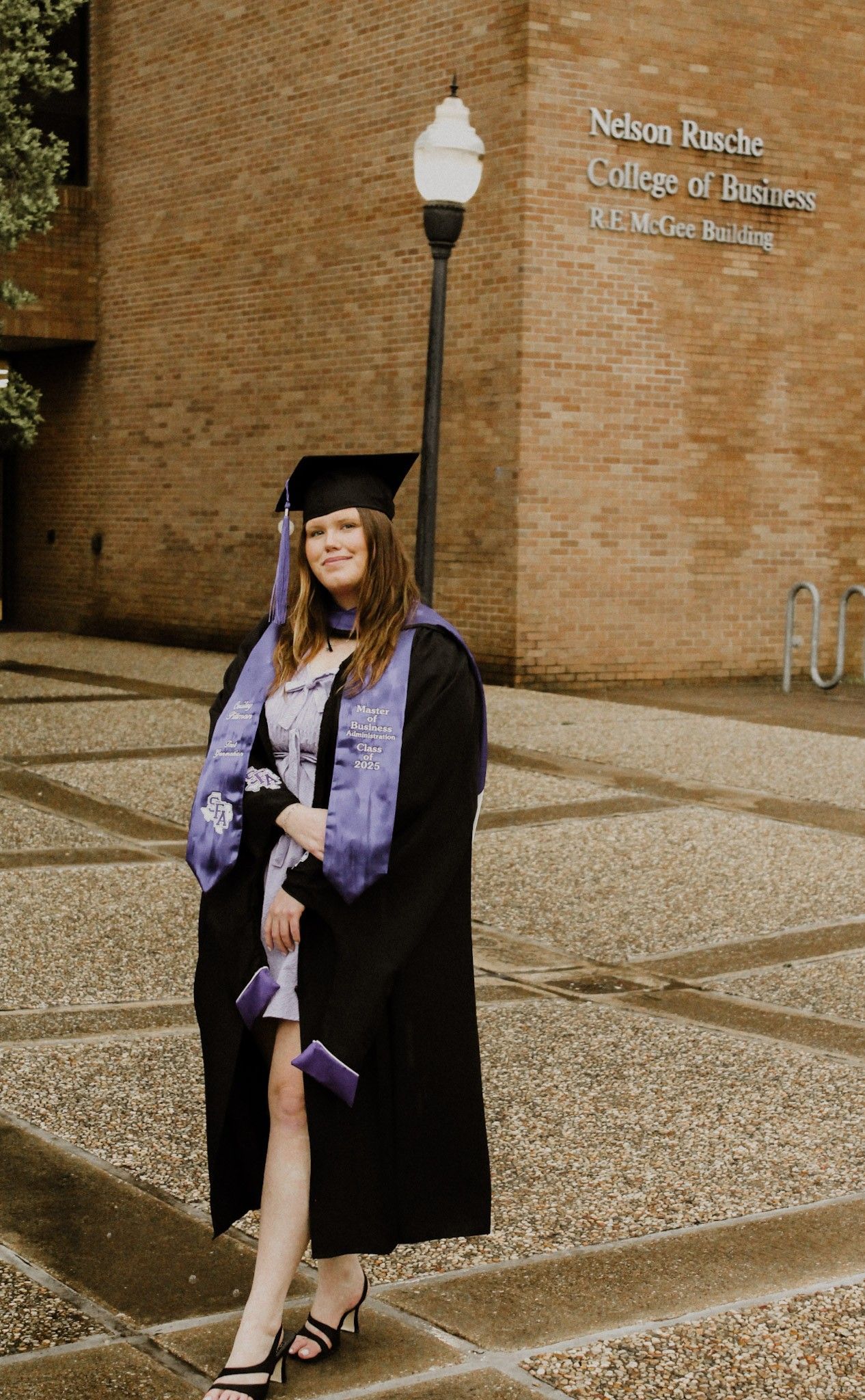 Woman in graduation attire stands in front of a brick building with a sign that reads 
