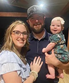 Family of three smiling, posing for a photo. A woman in glasses and a man holding a baby.