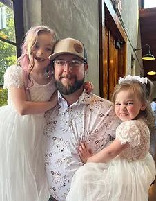 Man with two young girls, all smiling. Girls wear white dresses, man wears a hat and patterned shirt.
