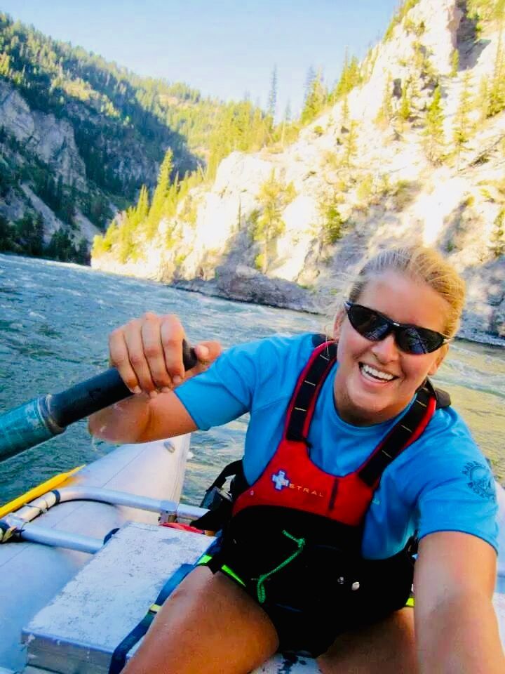 Woman wearing sunglasses and a life vest smiles while rowing a raft on a river, with mountains in the background.