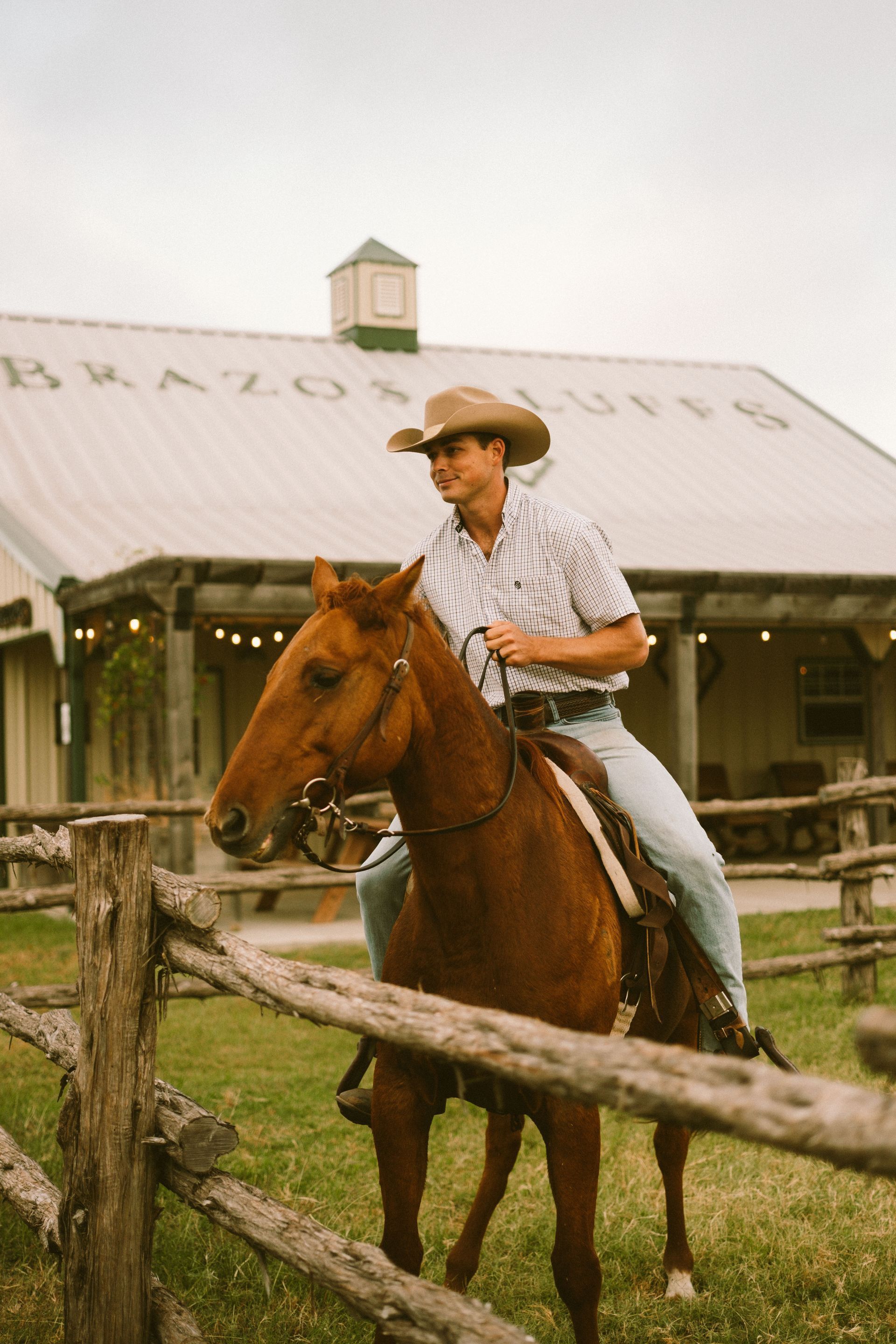 A person wearing a cowboy hat and western-style clothing rides a brown horse in front of a building labeled Brazos Bluffs.