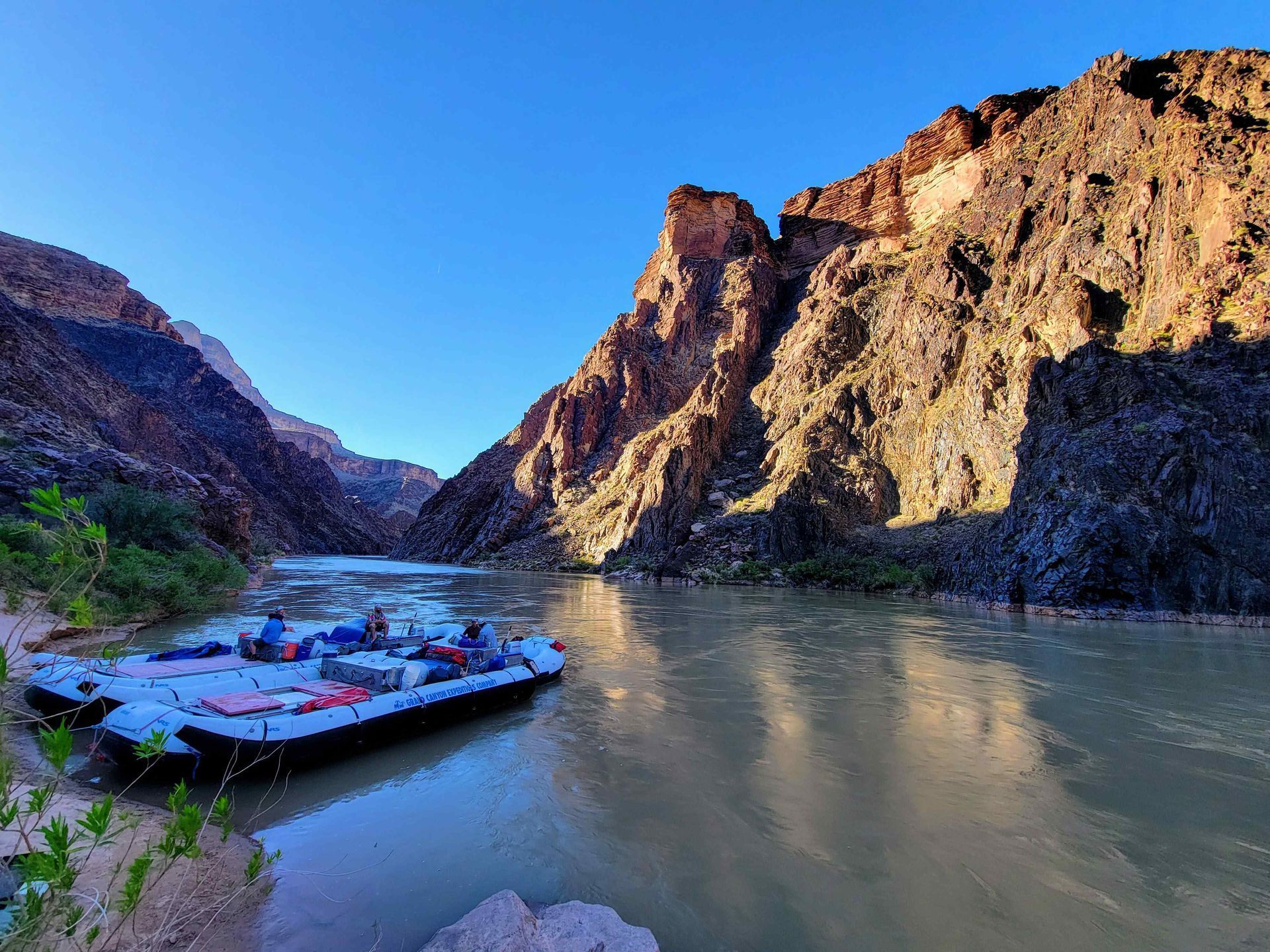 Rafts on a river in a canyon, bathed in sunlight. Dark rock cliffs rise above the water.