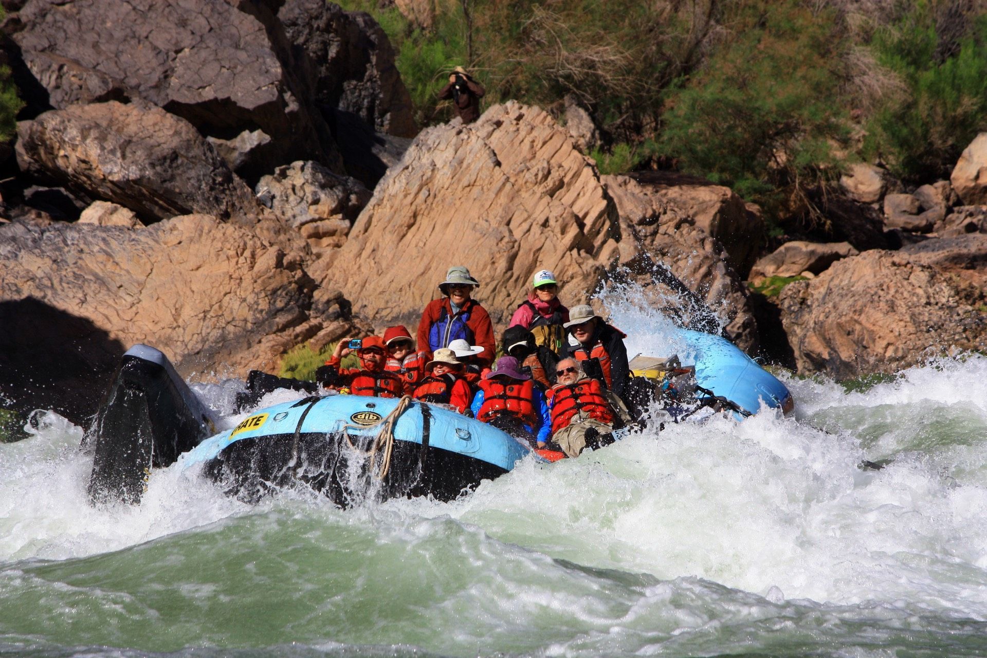 A group of people in life vests navigate a blue raft through whitewater rapids surrounded by rocky cliffs.