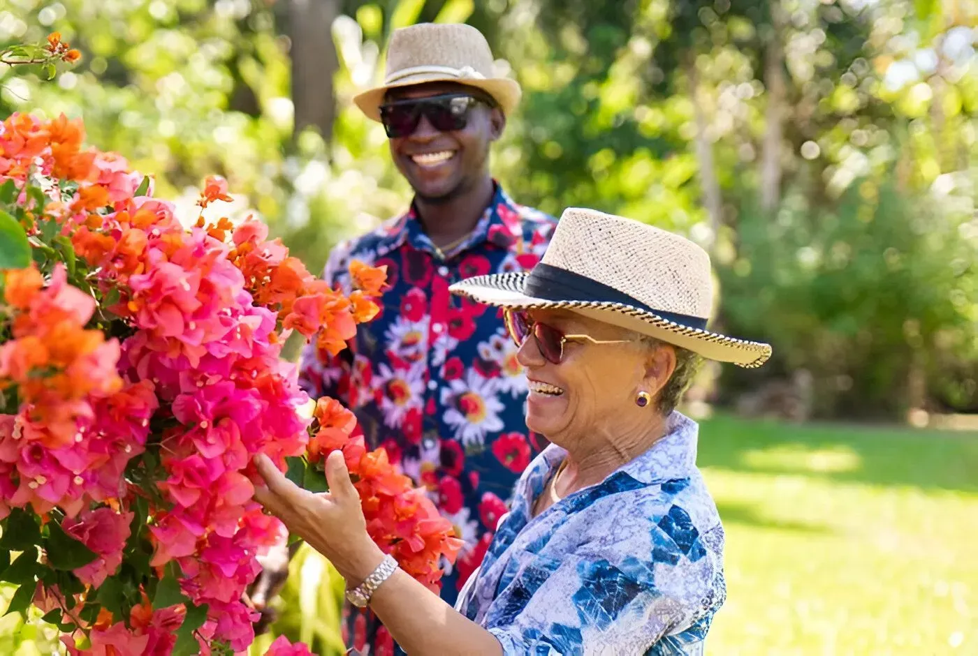 Woman touching bright pink flowers, man smiling in background, both wearing hats, outdoors.
