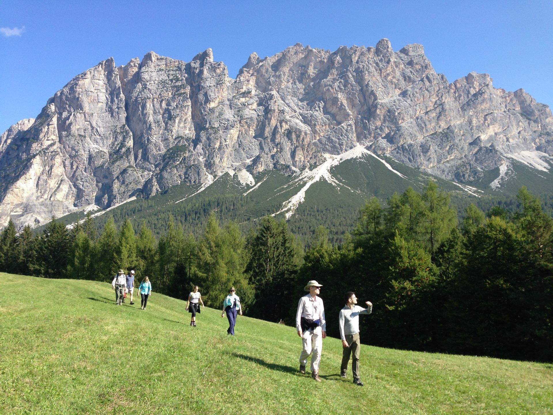 Hikers walk on a grassy hill toward a large, rocky mountain range under a clear blue sky.