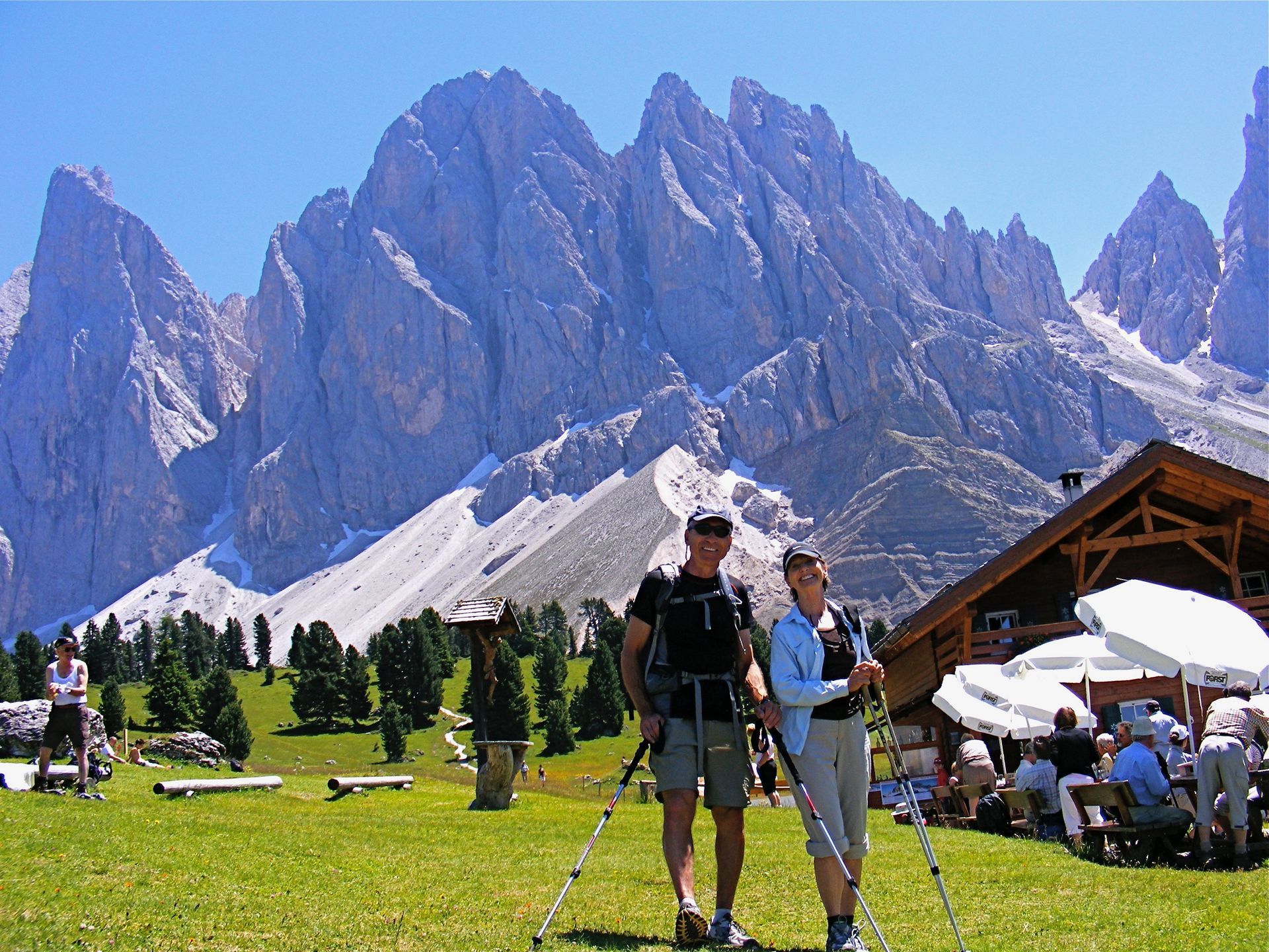 Two hikers with trekking poles pose in front of a mountain range. A wooden building and outdoor seating are nearby.