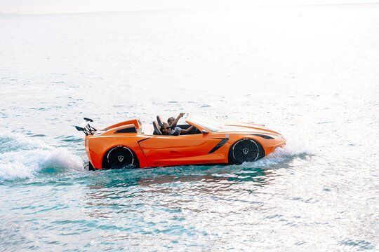 Orange convertible car driving on water, two people onboard waving arms in the ocean.