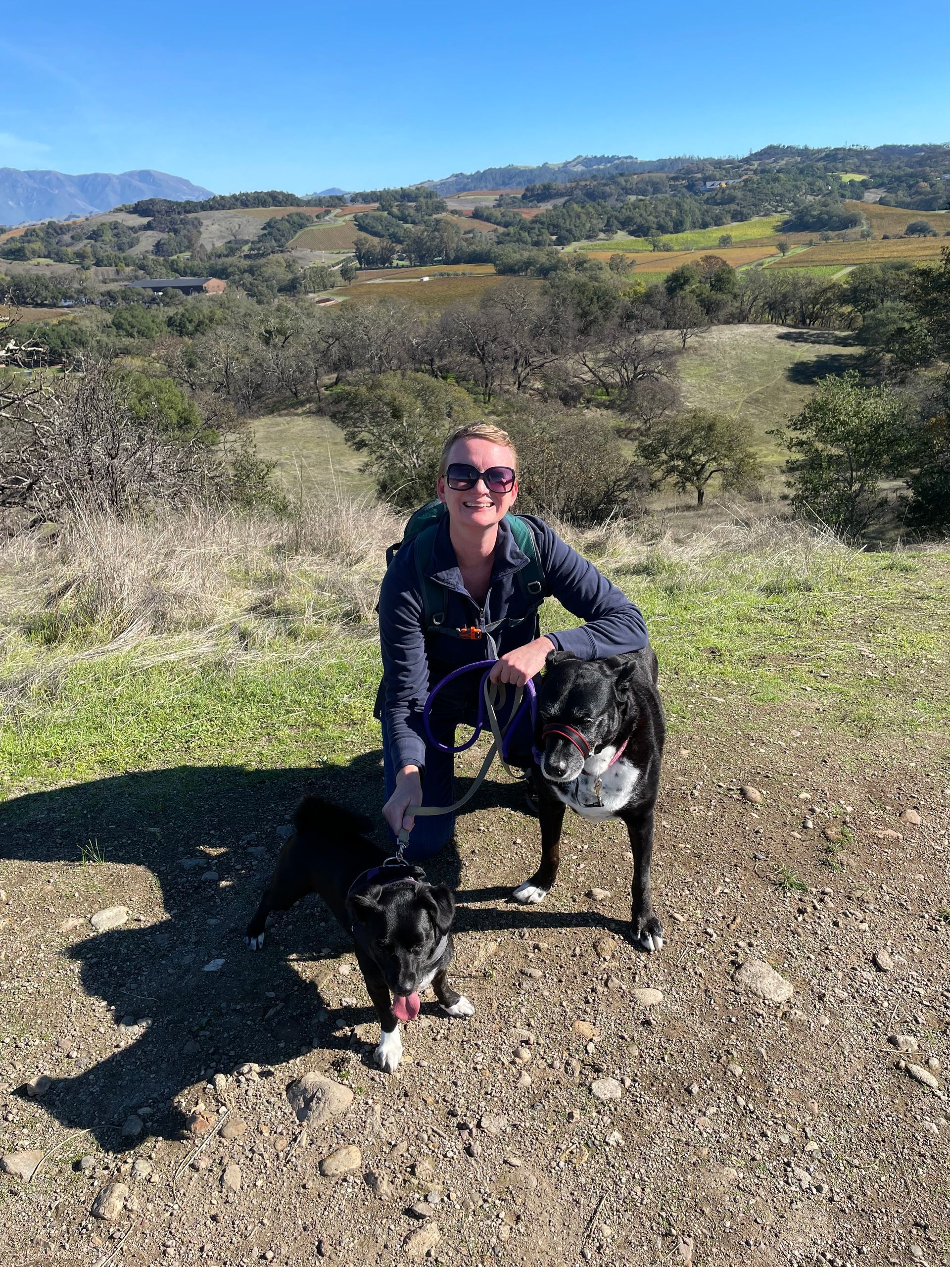 A woman is kneeling down next to a dog on top of a dirt hill.