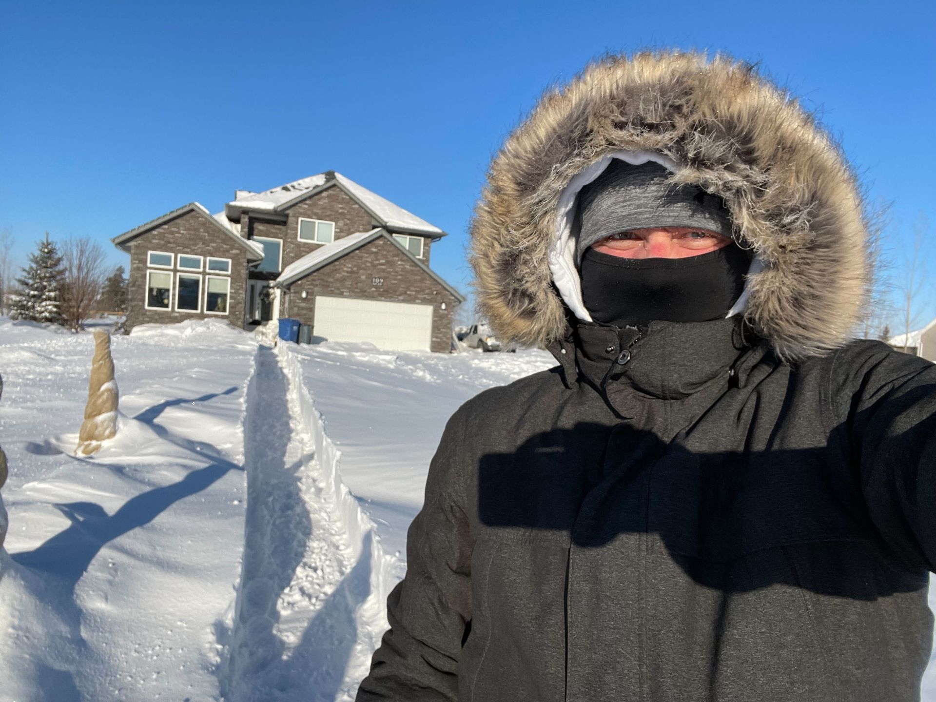 A man wearing a fur hooded jacket is taking a selfie in front of a snowy house