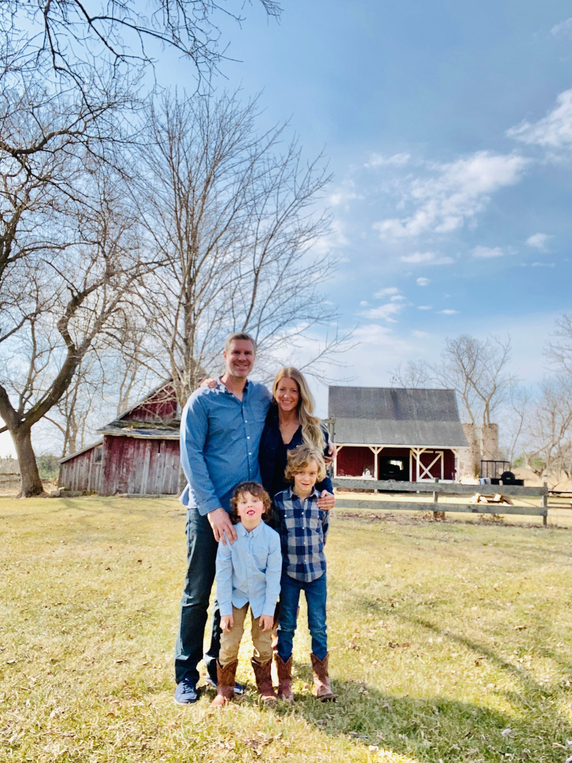 Family of four stands in a grassy field with a red barn in the background; sunny day with blue sky.