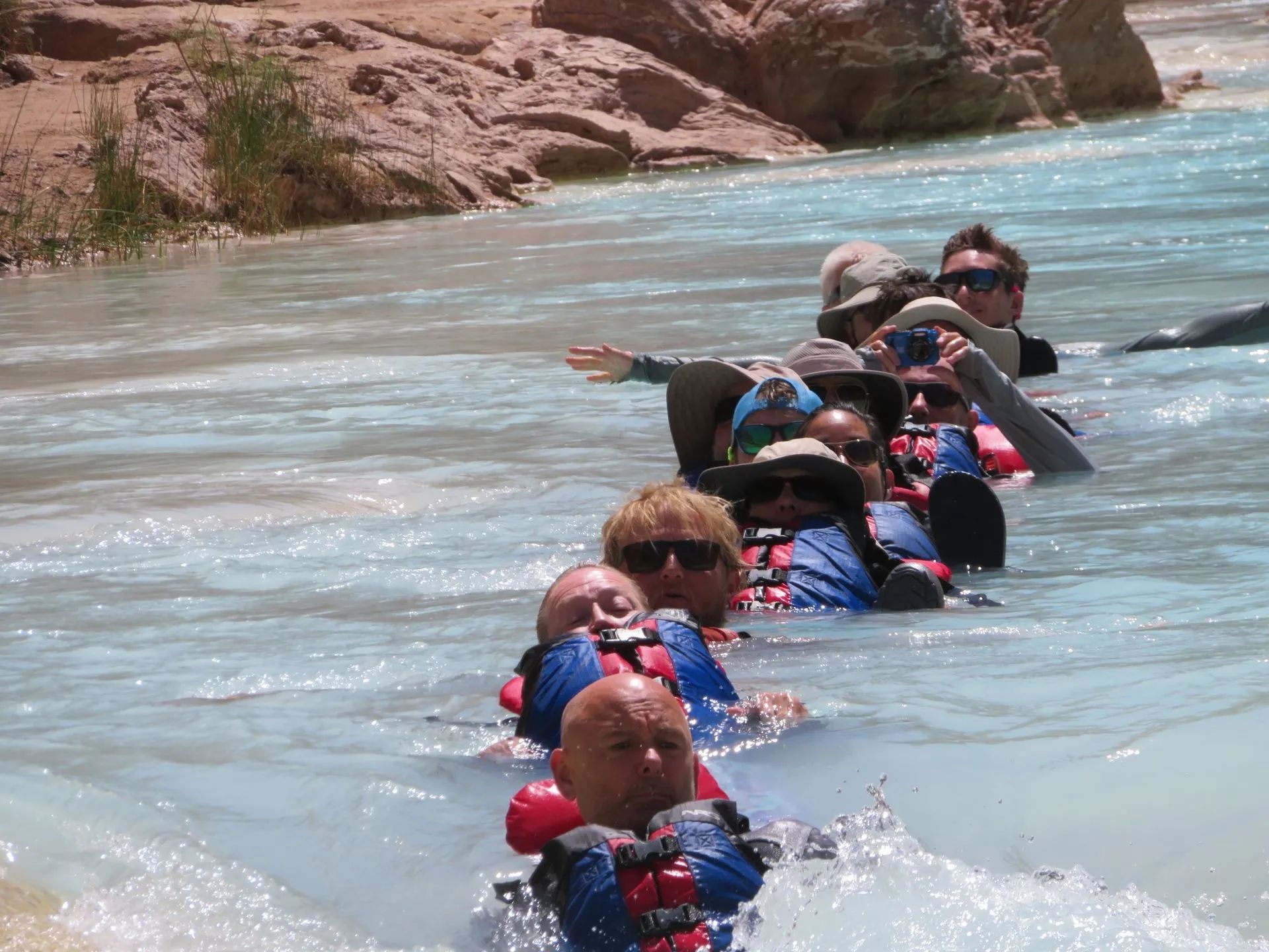 People wading through clear, blue-green water, wearing life vests. Red rock formations in background.