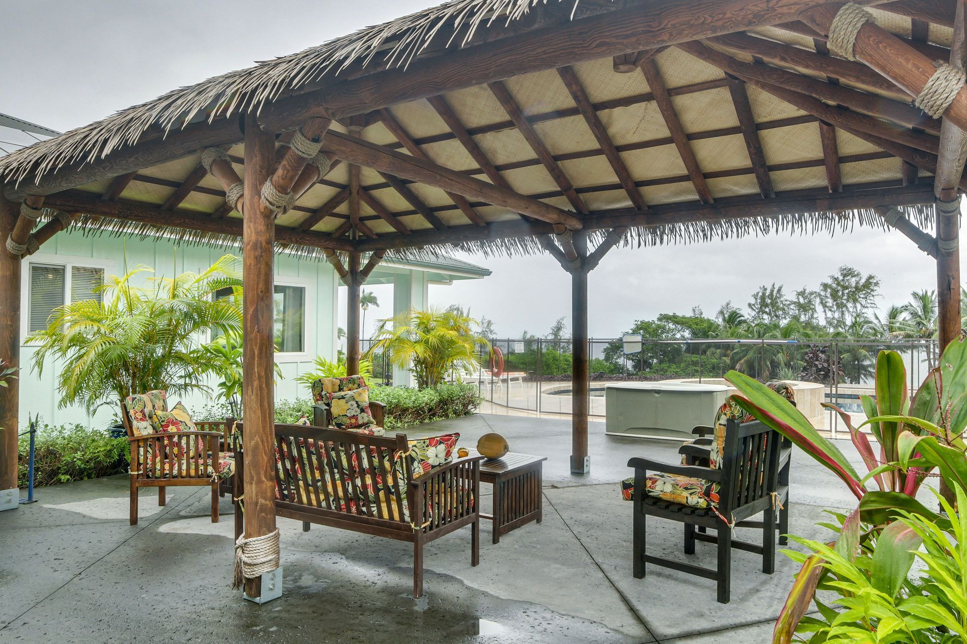 A shaded patio with wooden furniture and a thatched roof. Green foliage and a light blue building are visible.