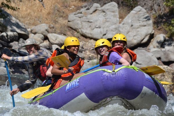People on a purple raft white-water rafting. They wear yellow helmets and life vests. Rocky background.