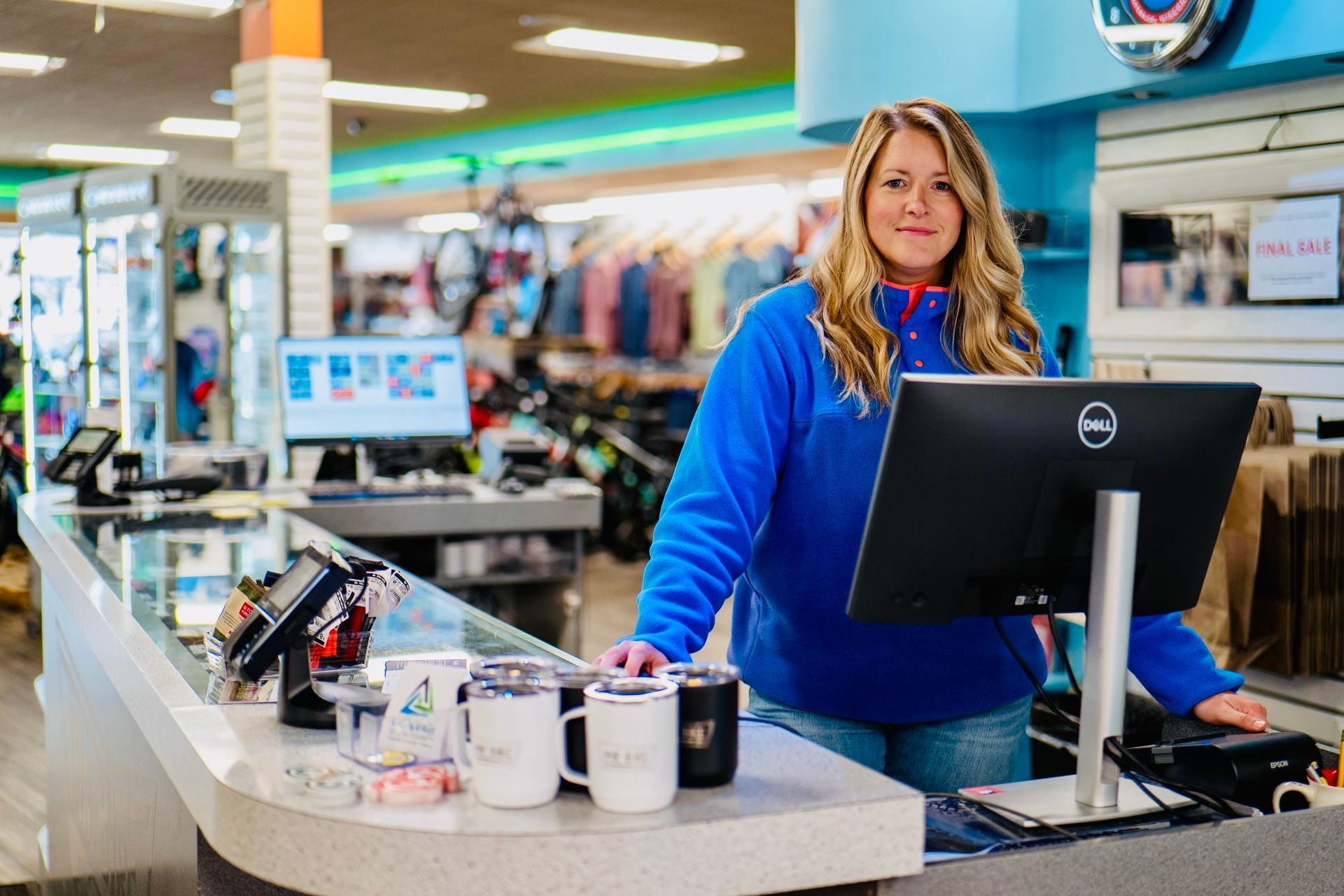 Woman behind a store counter, wearing blue fleece, smiling. The store has blue walls and sells merchandise.