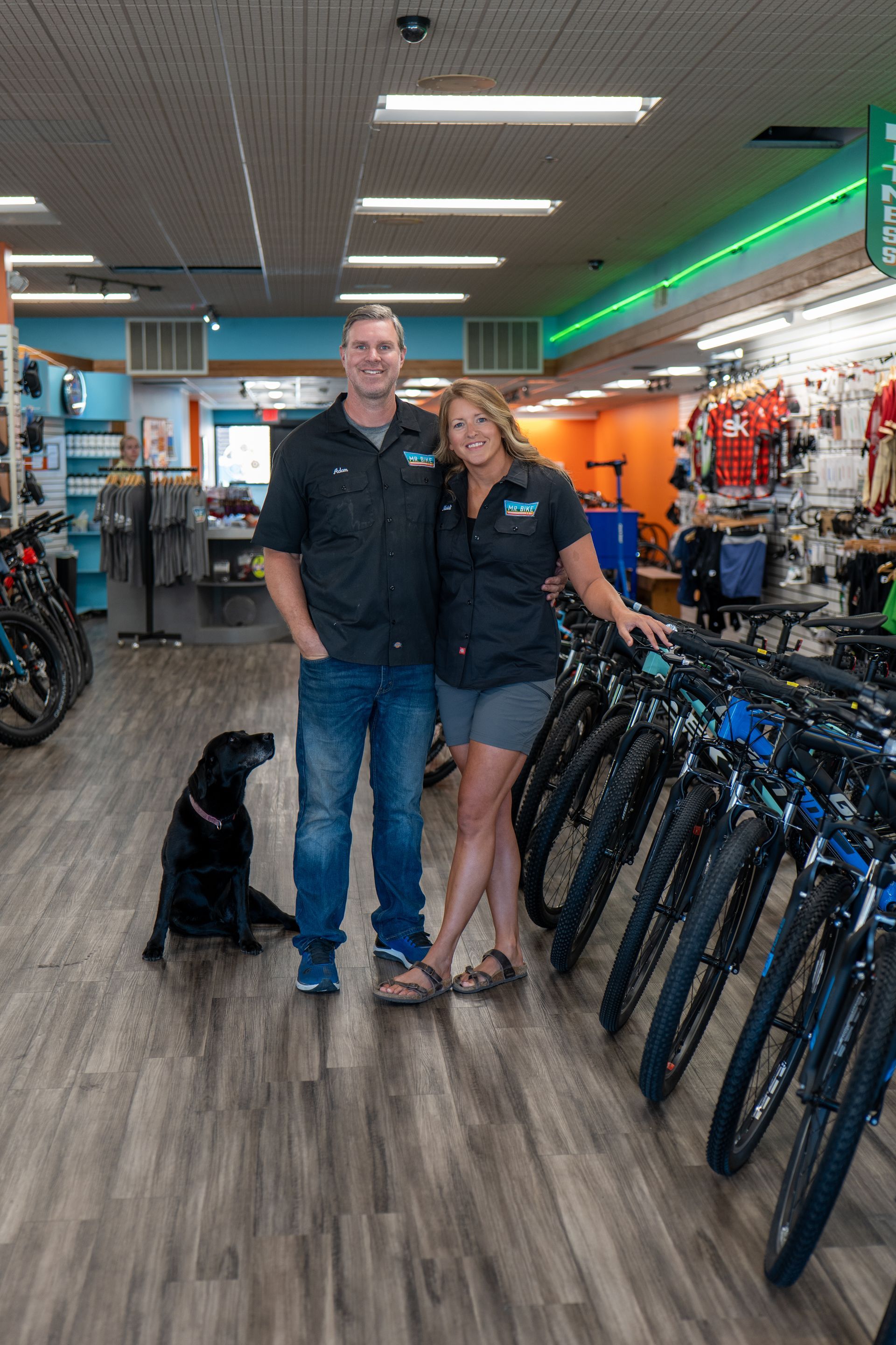 Man and woman standing in a bike shop with a black dog. They are posing next to a row of bicycles.