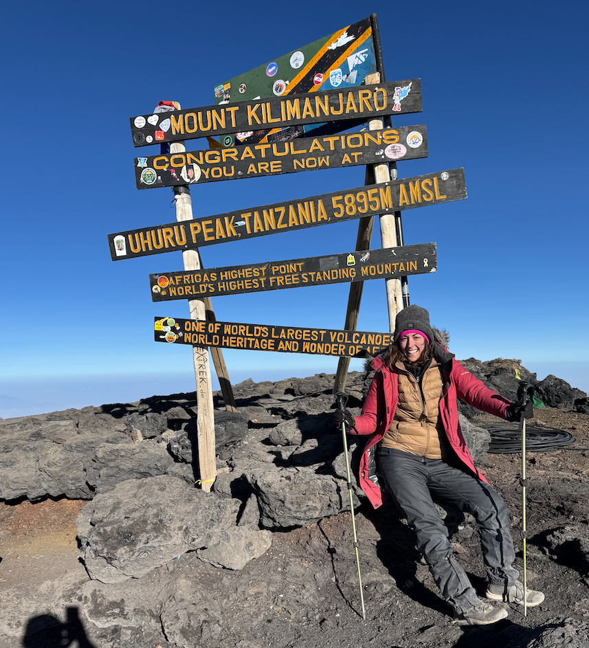 Woman at Mount Kilimanjaro summit sign, smiling, red jacket, blue sky.