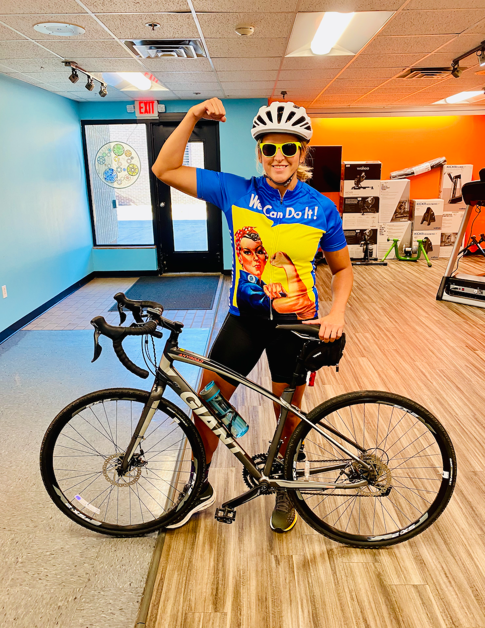 Woman in cycling gear flexes, posing with a bike inside a store. She wears a helmet, sunglasses, and a blue jersey.