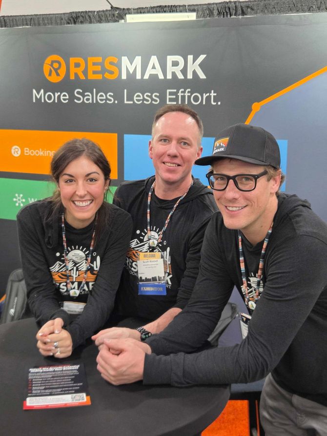 Three people smiling at a RESMARK booth, seated in black shirts with orange and gray signage behind them.