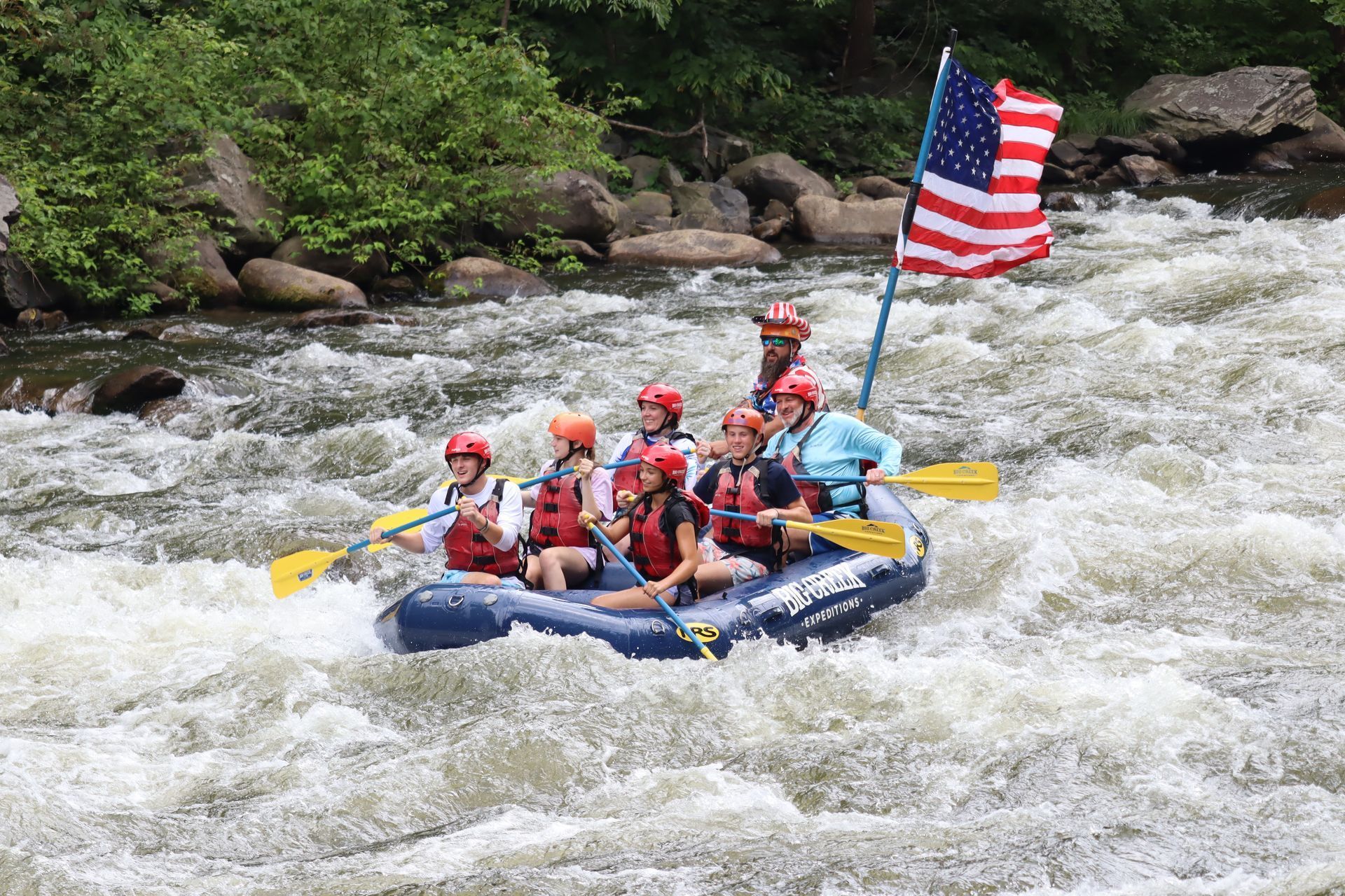 Whitewater raft with helmeted paddlers and an American flag rushing through rapids