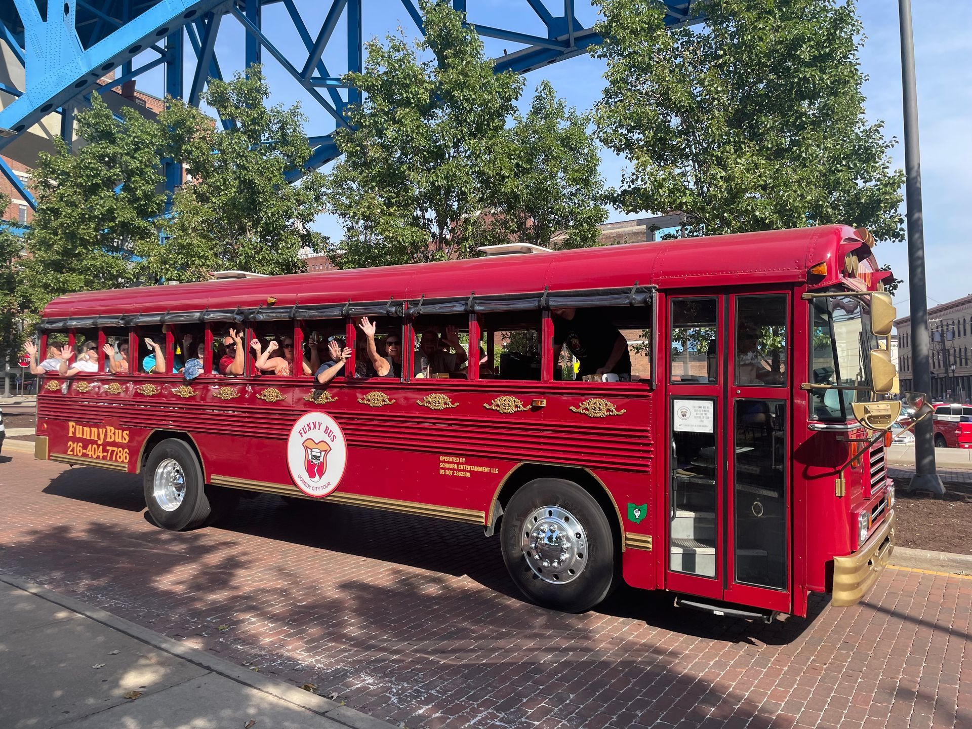 Red open-air trolley bus with passengers, parked beside a blue steel bridge on a city street.